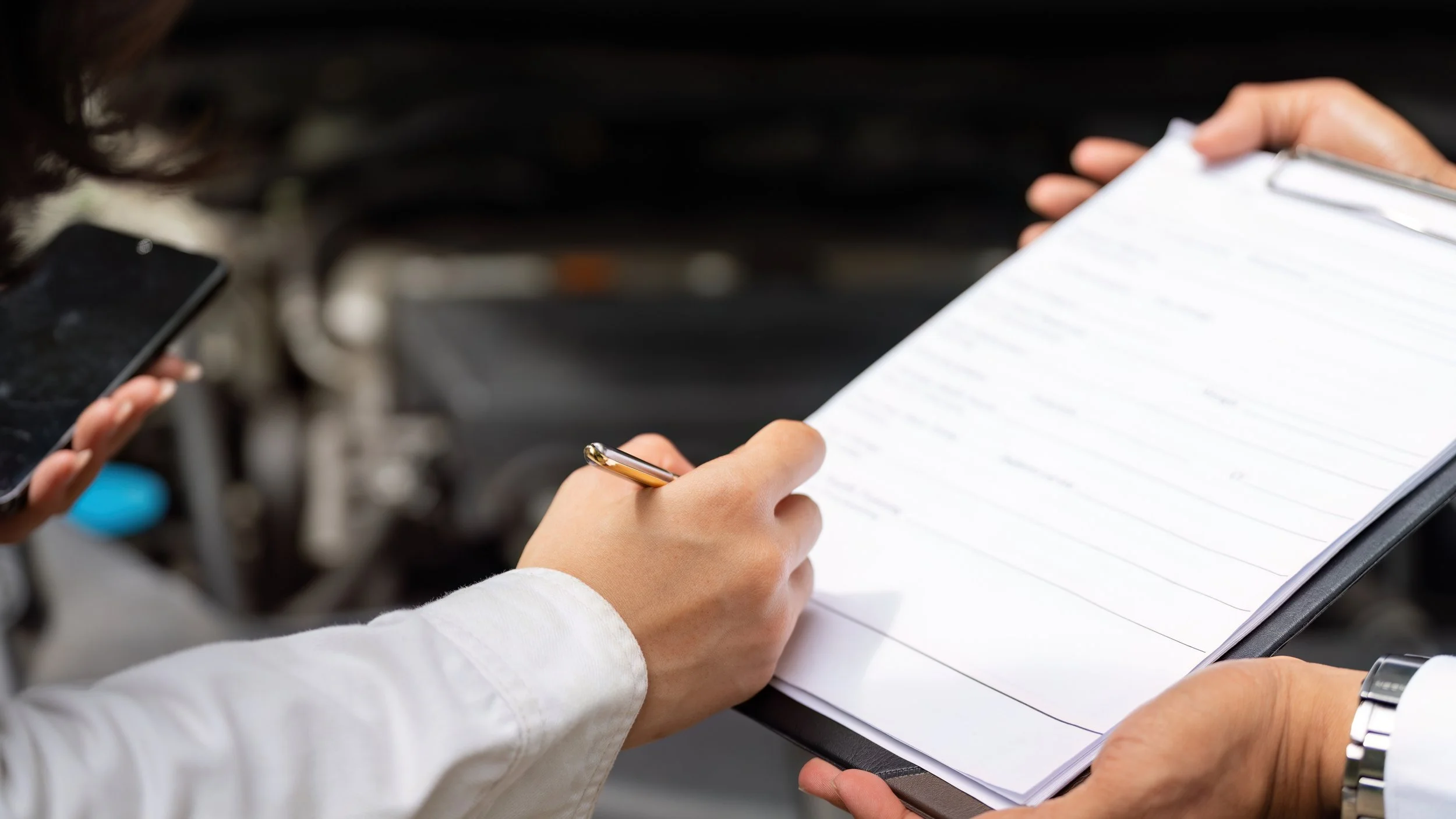 Person holding a clipboard with documents, writing with a pen, and another person holding a smartphone nearby.