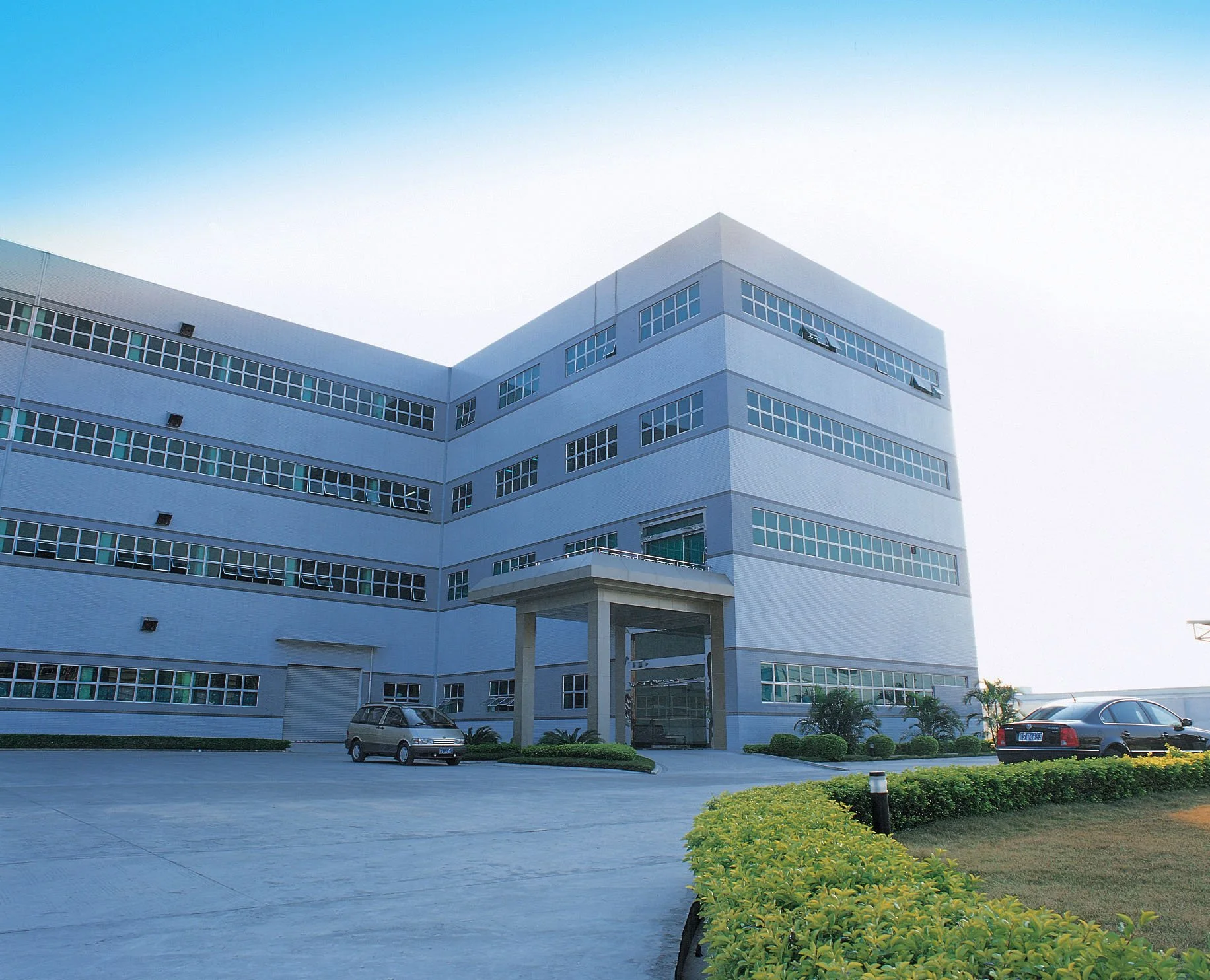 Modern multi-story office building with large windows, parked cars in front, landscaped greenery, and a clear sky.