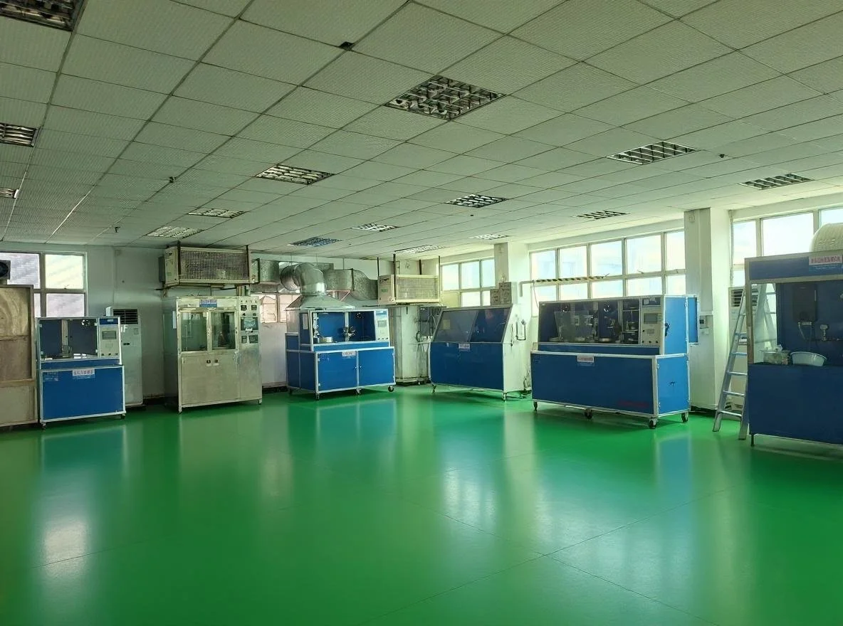 Laboratory with multiple blue and silver scientific instruments and equipment aligned along the wall, bright natural light from large windows, green floor, and ceiling with square tiles.