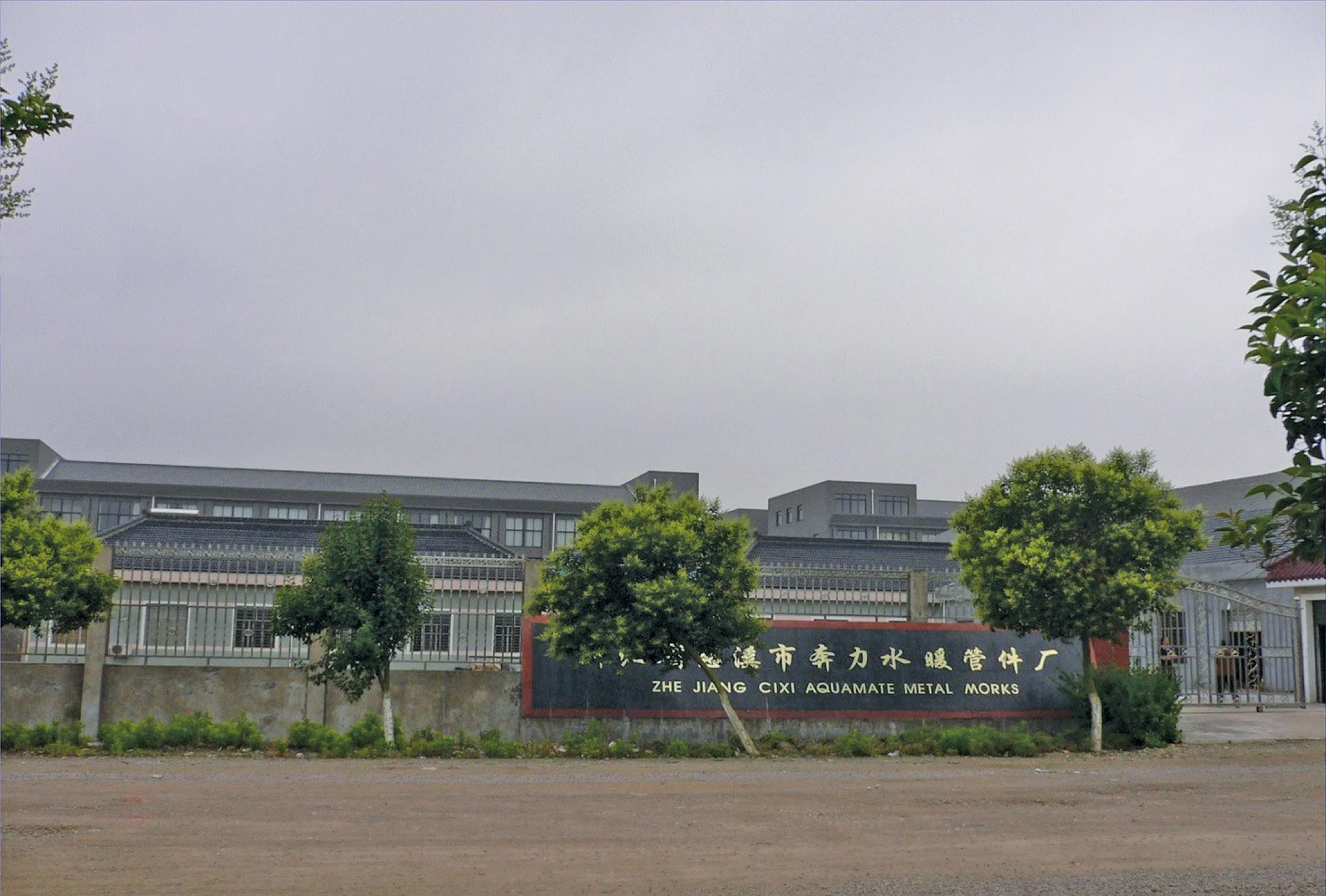 A factory building with a black sign in front reading 'ZHE JIANG CIXI AQUAMATE METAL MORES' surrounded by trees and a dirt road