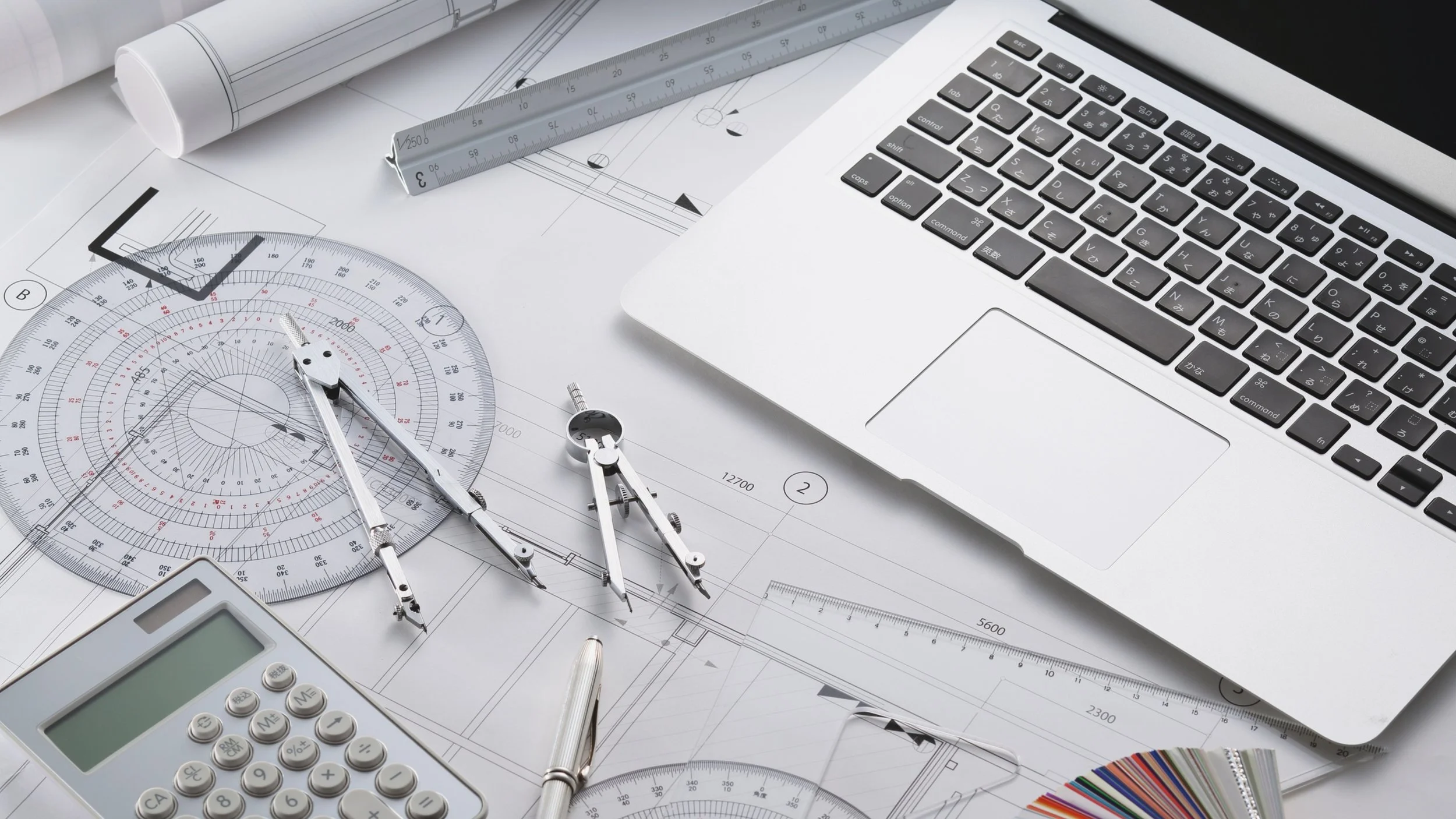 Architectural drawing tools including a protractor, compass, ruler, a calculator, a pen, and a computer keyboard on blueprints.