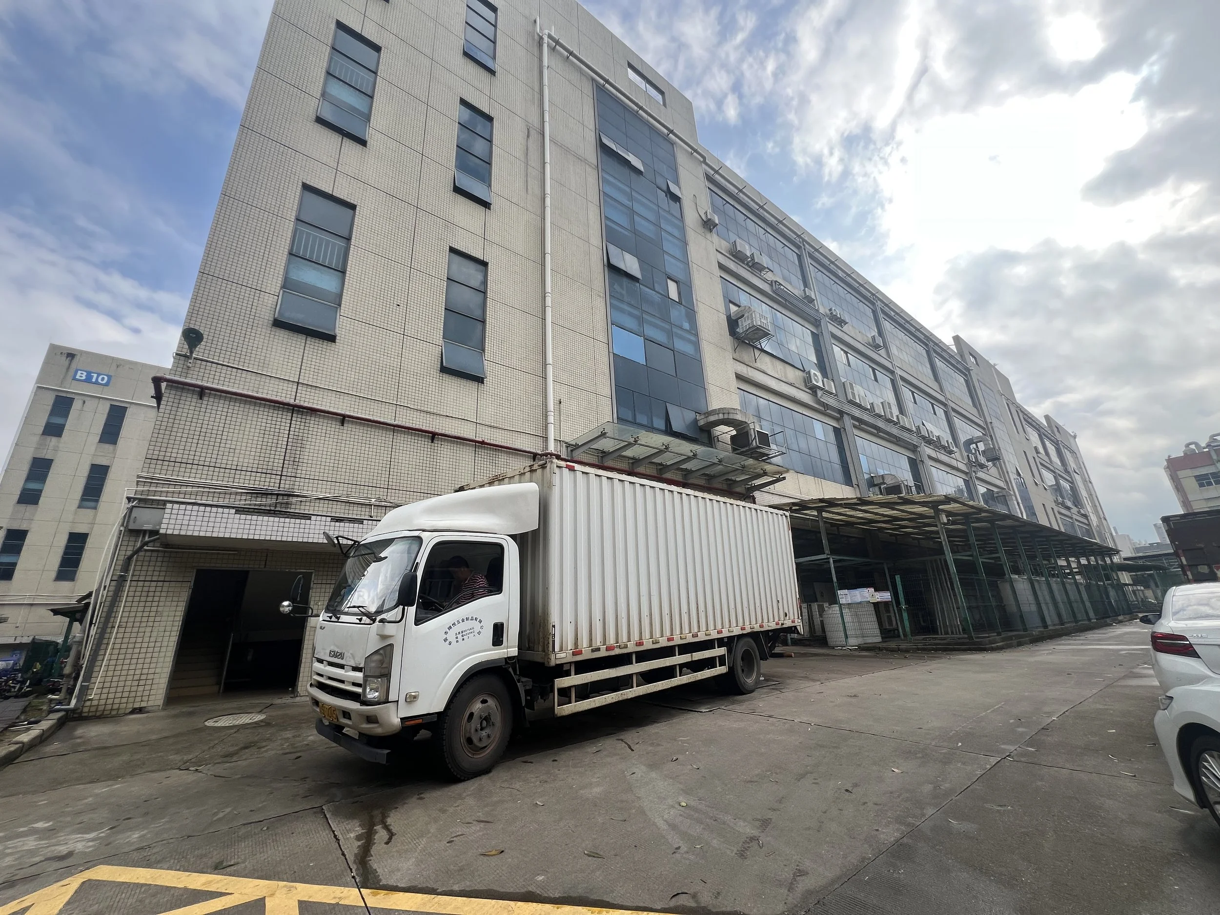 A white delivery truck parked in front of a multi-story office building with glass windows and air conditioning units, under a cloudy sky.