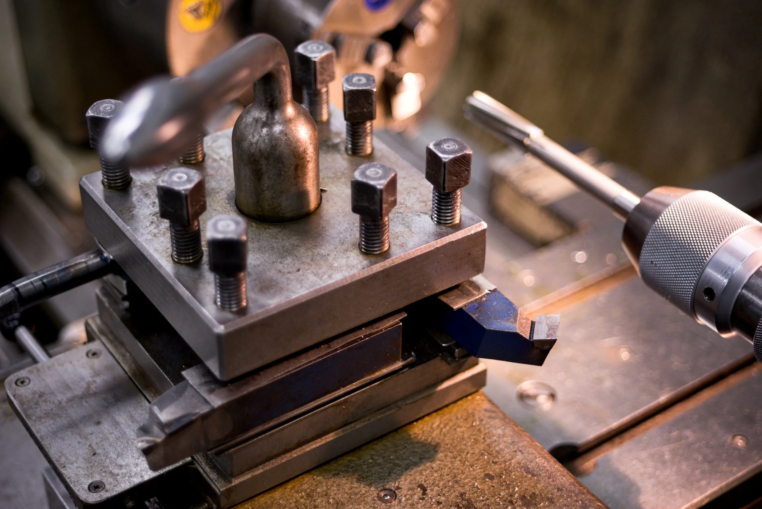 Close-up of a metalworking machine with a metal piece clamped in place, surrounded by bolts, and a portable drill resting nearby.