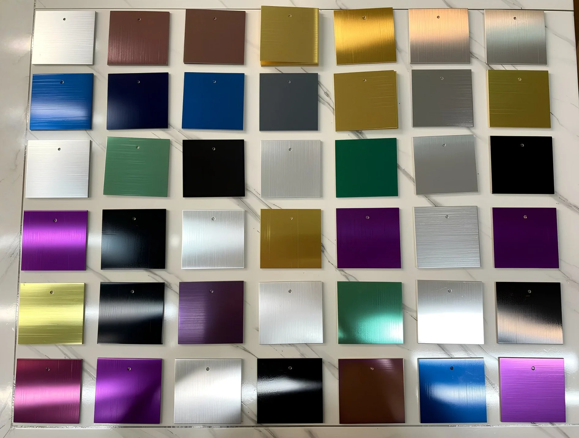 A display of square color samples arranged in a grid pattern on a white surface. The samples include a variety of colors such as gold, black, blue, green, purple, gray, silver, maroon, and white, with some featuring a metallic or brushed texture.