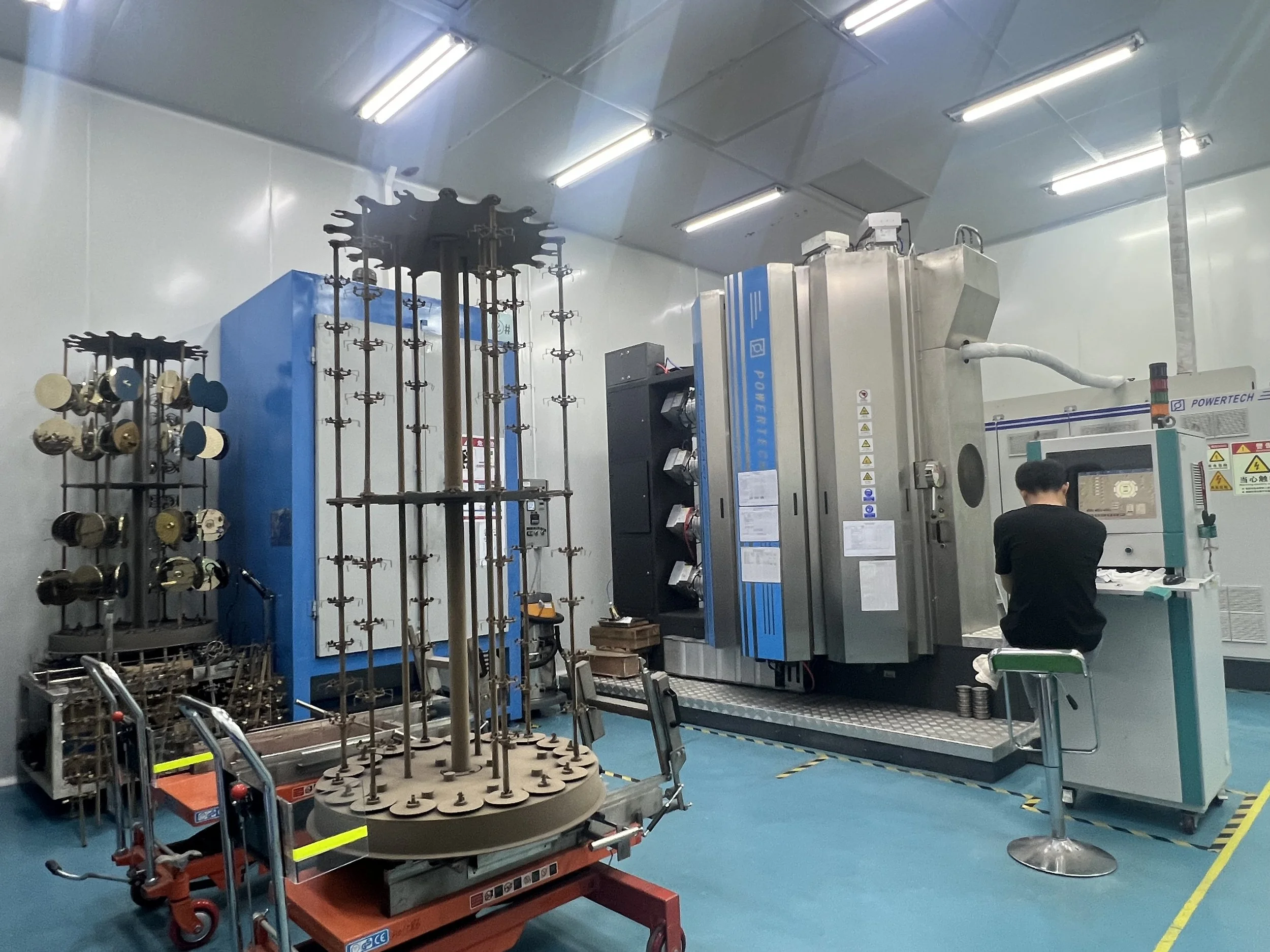 An engineer sits at a control panel of a PVD coating machine in a laboratory with scientific equipment and machinery, including a large blue machine with multiple components and a circular metal structure with rods and fixtures.