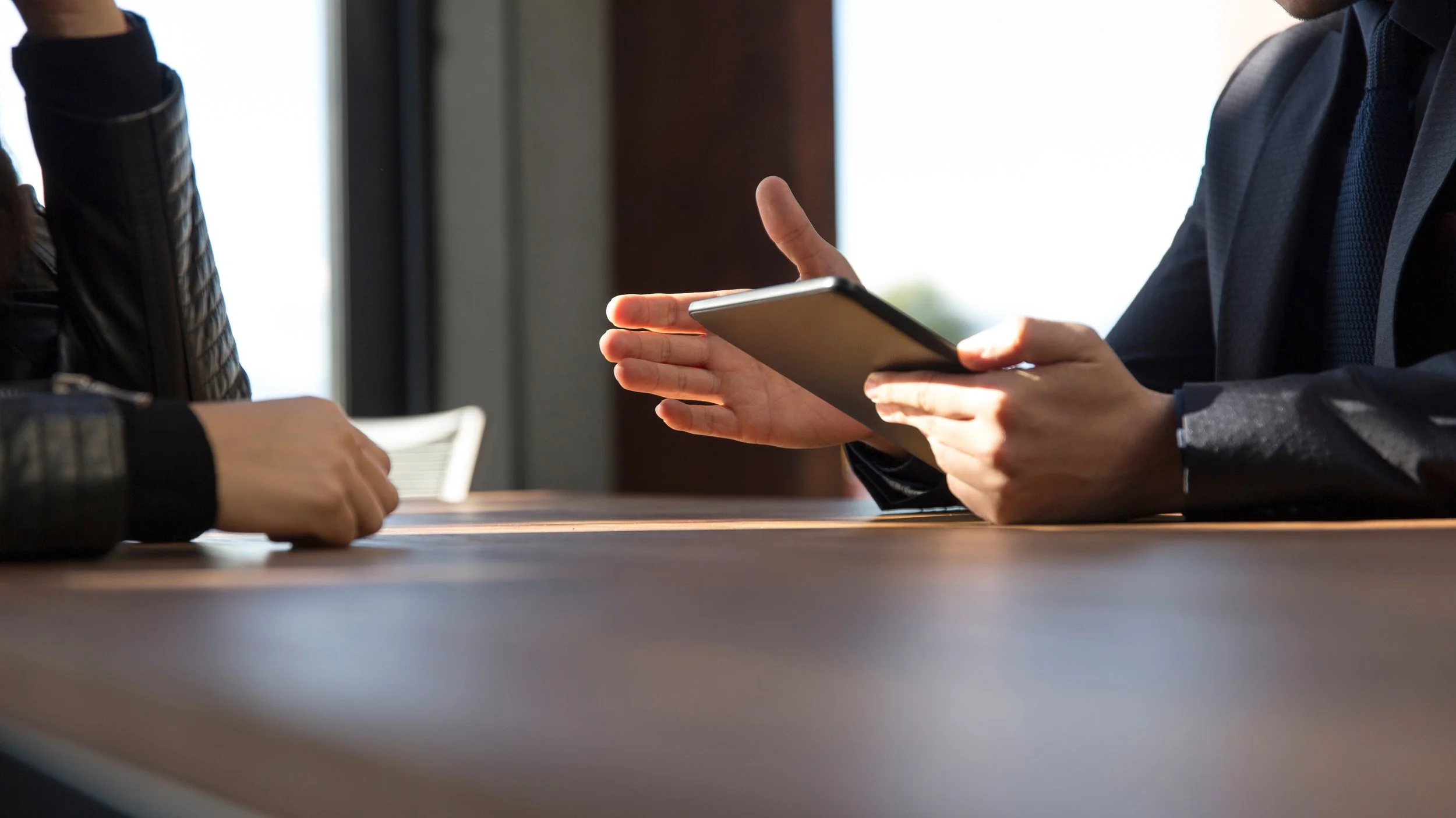Two people sitting at a table, engaged in a conversation. One person is holding a tablet, gesturing with their hand. The other person's hands are resting on the table.