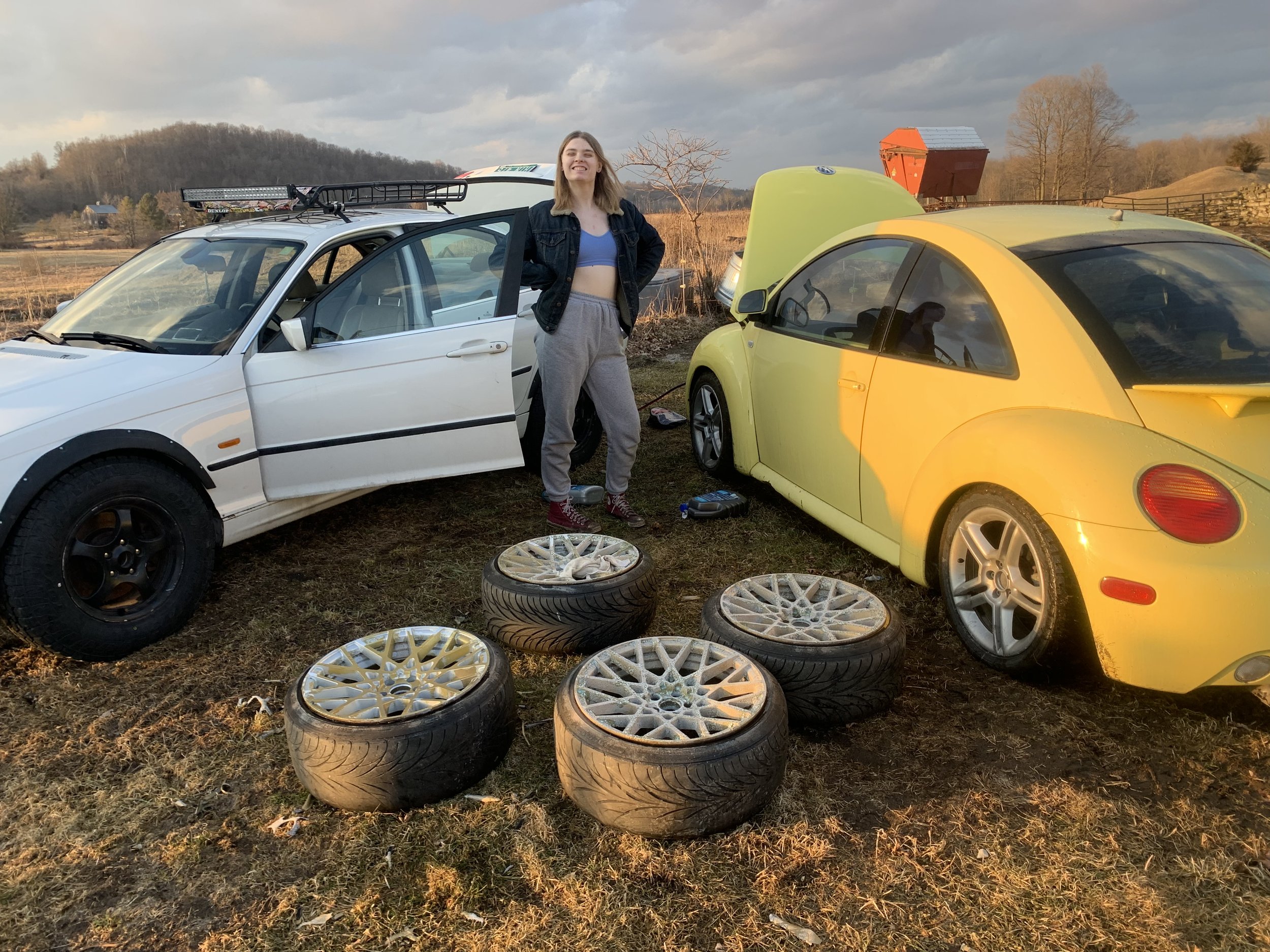 A young woman standing between two cars, a white sedan with a roof rack and a yellow sporty car with an open hood, with four tires and rims on the ground in front of her, outdoors during sunset.