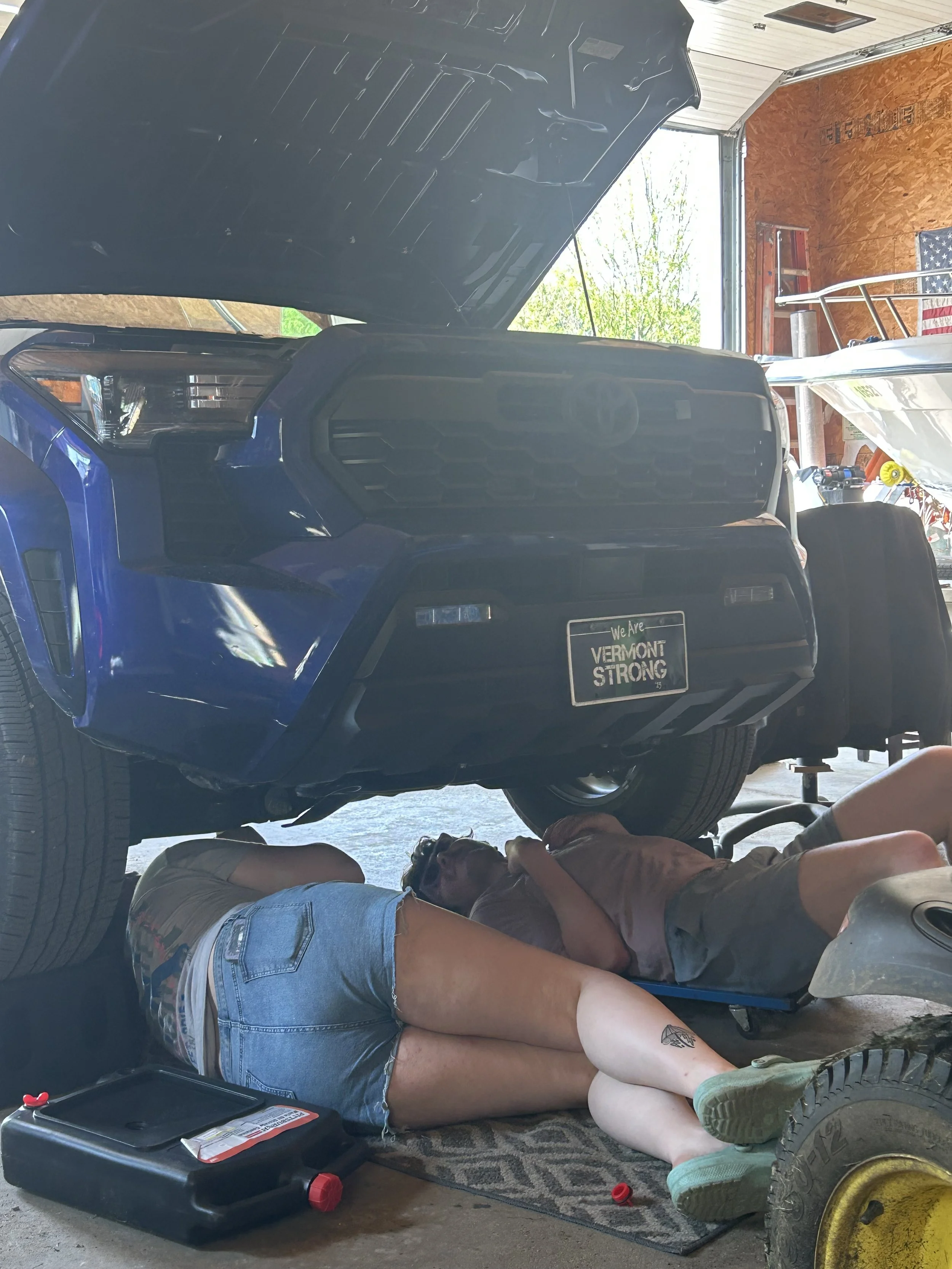 Two people lying underneath a blue truck in a garage, working on the vehicle's underside. The garage has wooden walls, tools, and an American flag hanging.
