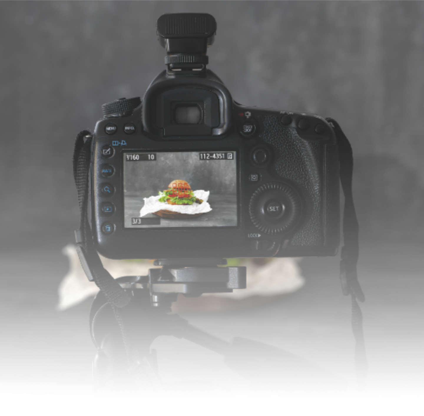 A digital camera on a tripod capturing a photo of a sandwich with lettuce, tomato, and other ingredients on a white paper towel, with a neutral gray background.