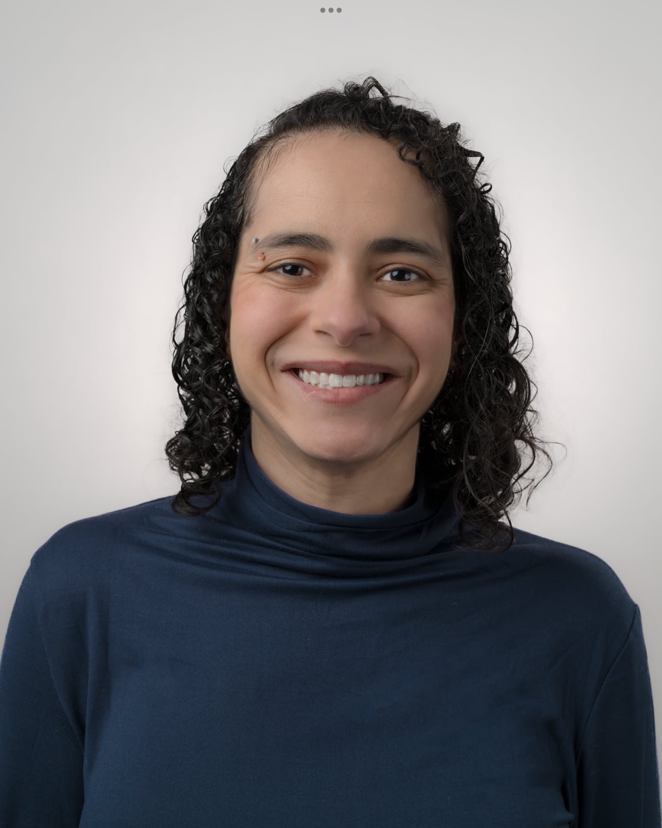 Person with curly dark hair and a blue turtleneck smiles at the camera against a plain light gray background.