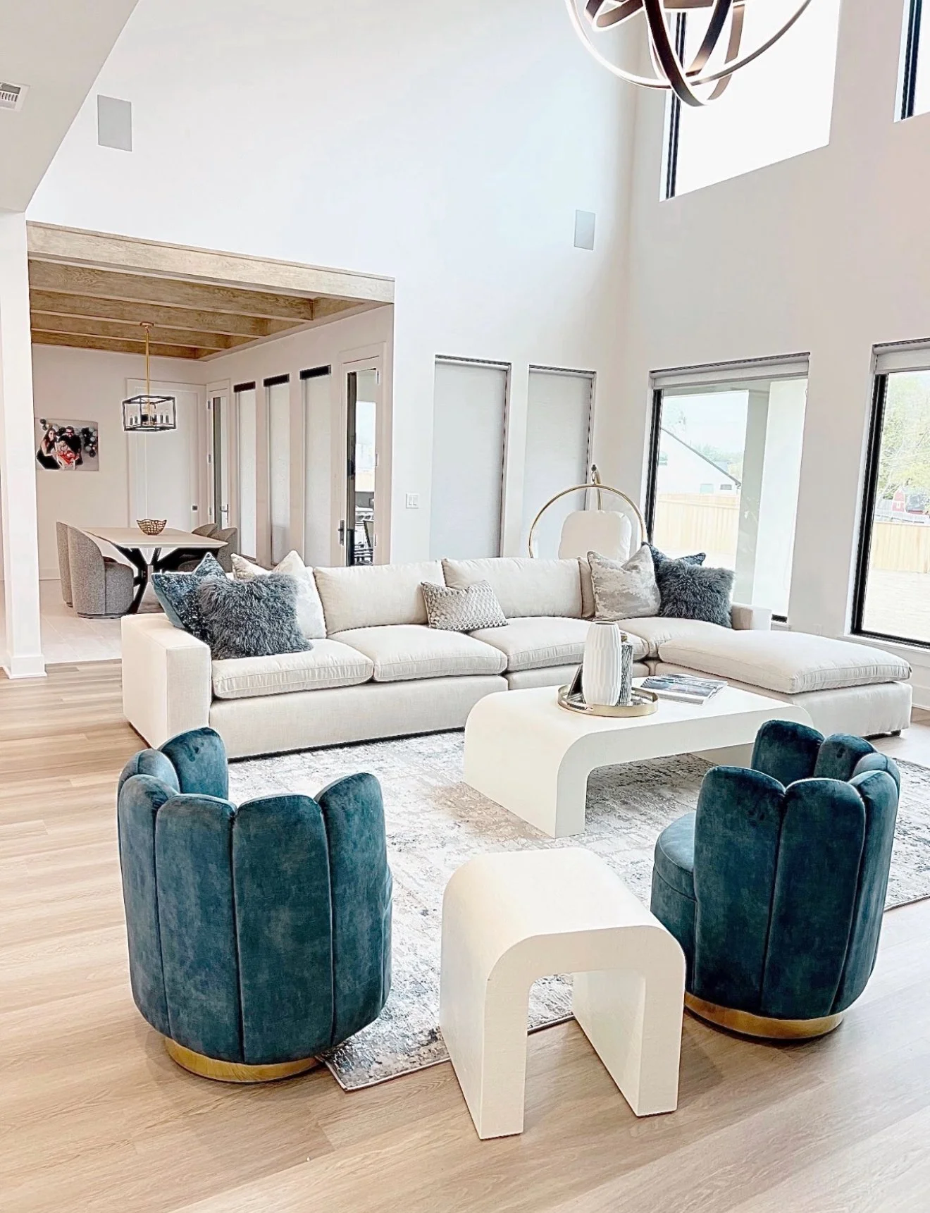 Bright living room with a large white sectional sofa, two blue velvet armchairs, and modern white coffee tables. The room has high ceilings, large windows, and a wooden ceiling in the dining area visible in the background.