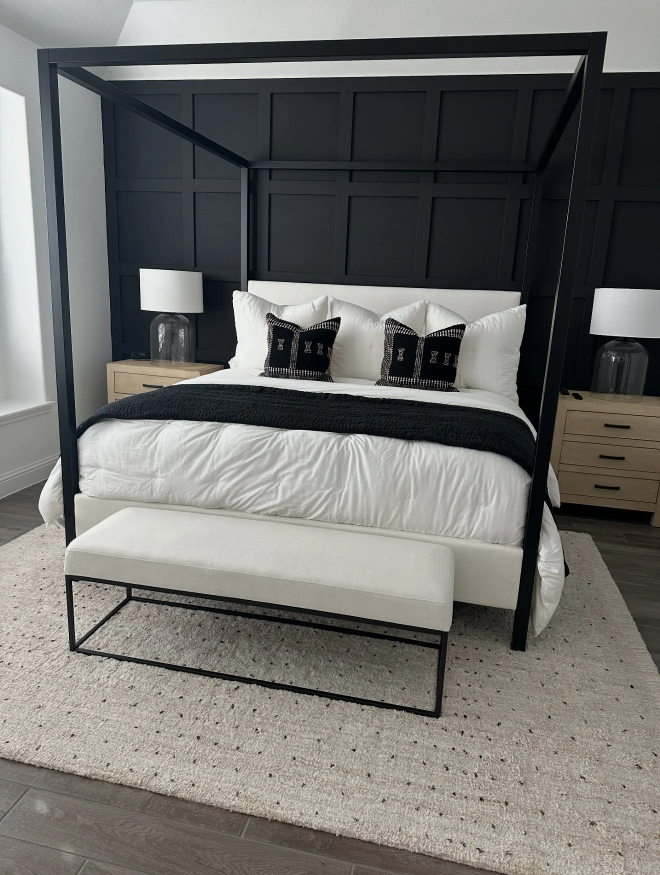 A modern bedroom with a white bed, black and white pillows, a black canopy frame, two wooden nightstands with glass lamps, and a white bench at the foot of the bed on a beige rug.