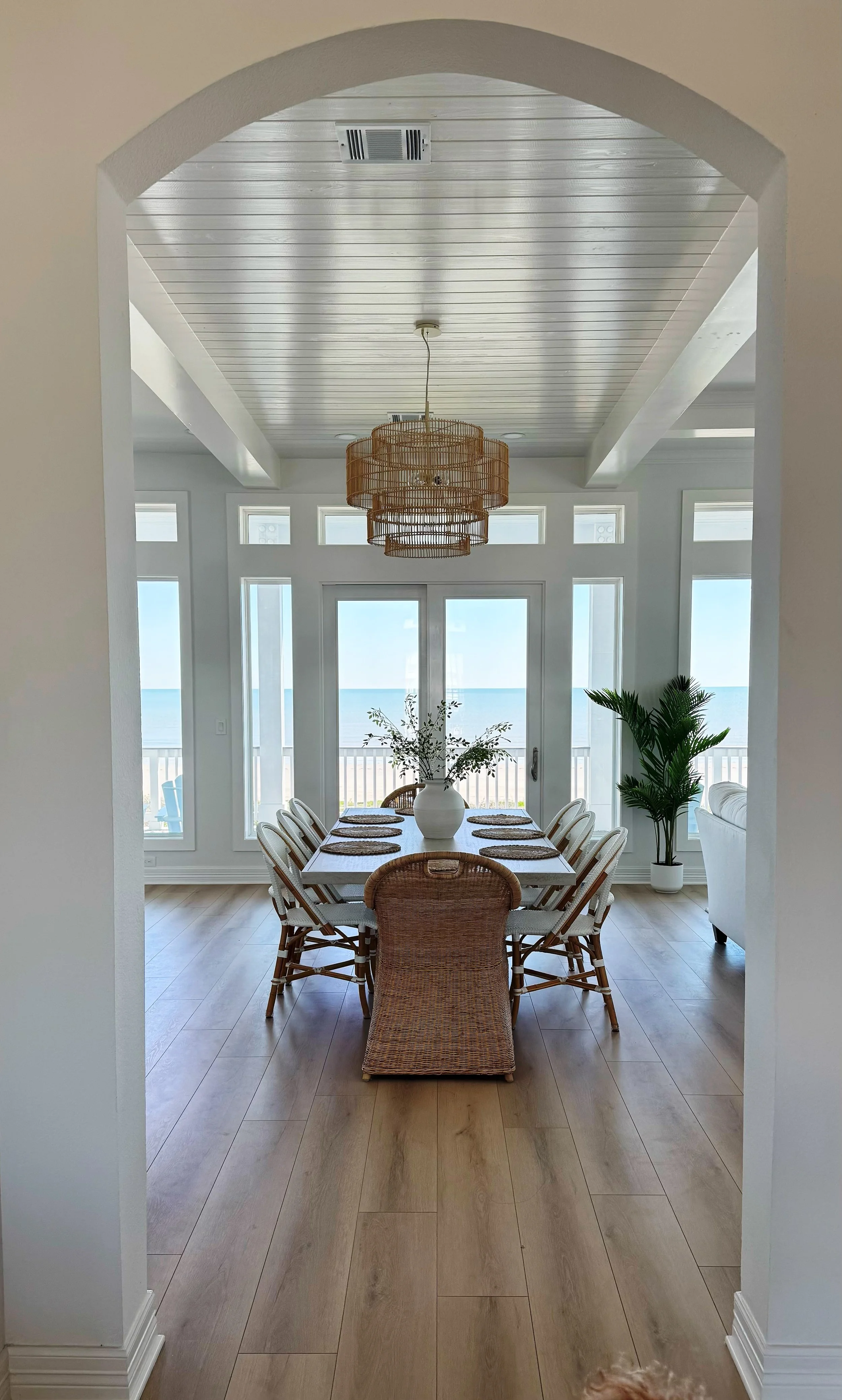 Dining area with a long table, ten chairs, a vase with greenery, large windows, and a view of the ocean.