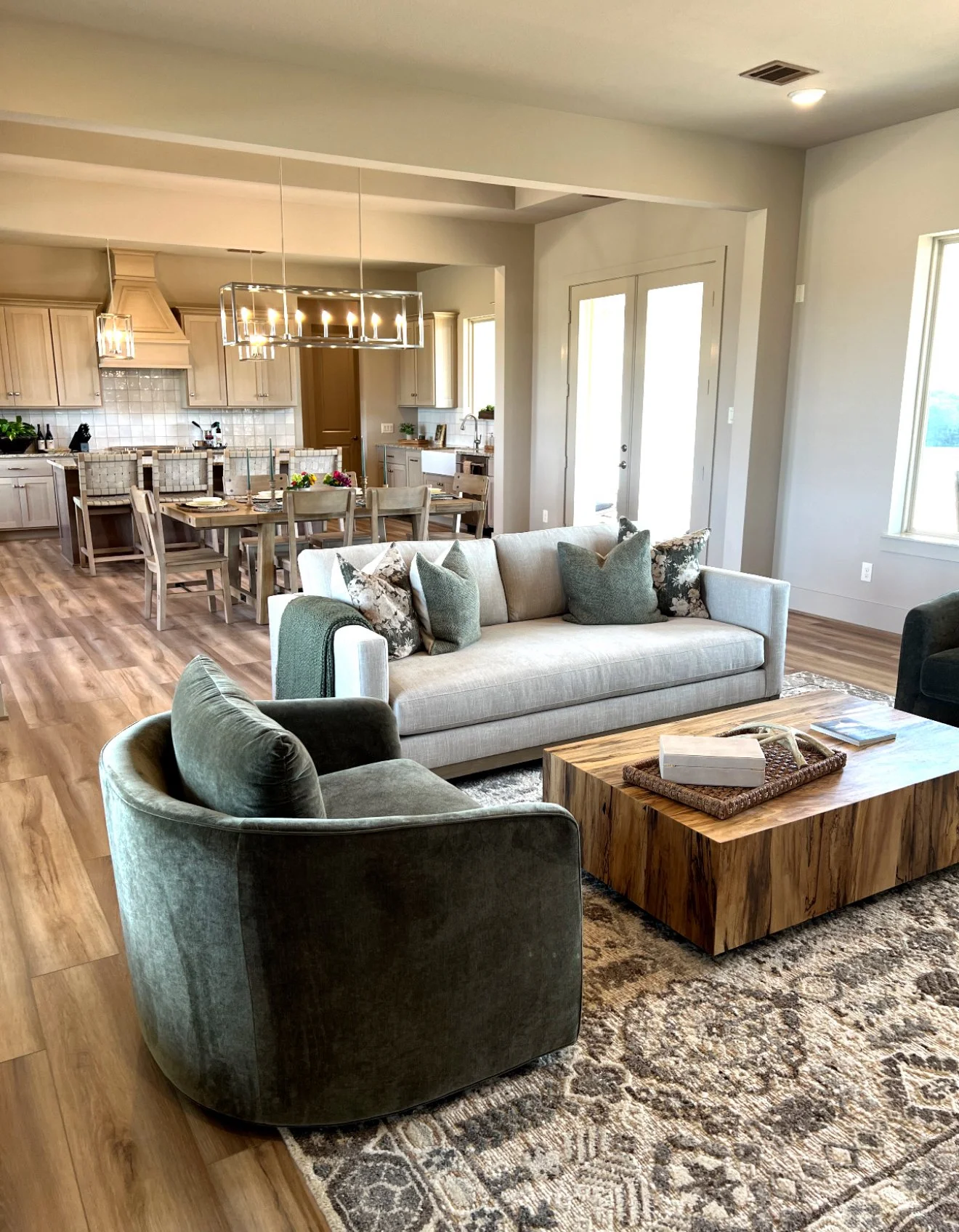 Living room with beige sofa, green armchair, wooden coffee table, and patterned rug, open to a dining area and kitchen with light wood cabinetry.