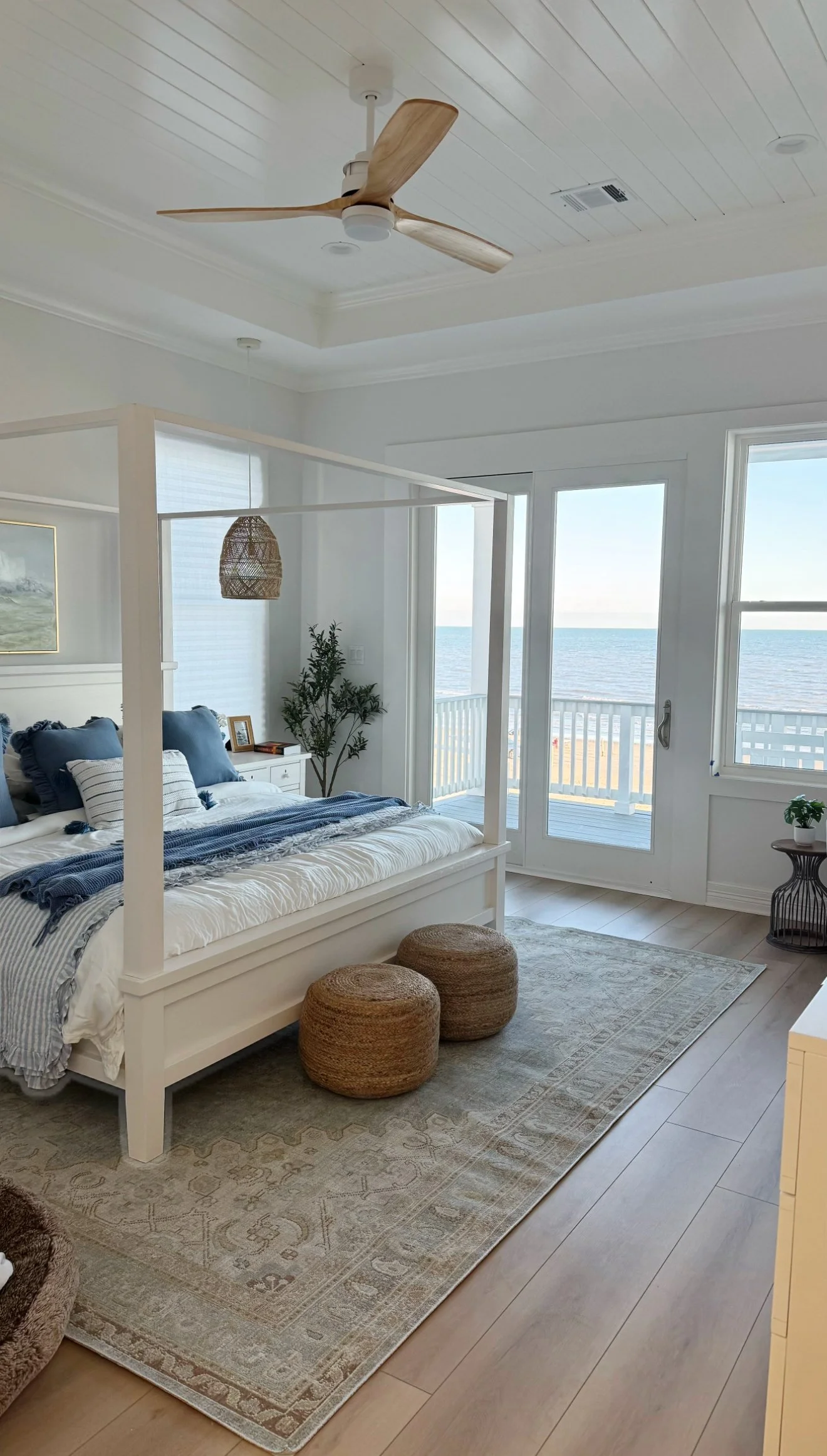 A bedroom with a white four-poster bed, decorated in blue and white bedding, in front of large windows showing a beach view. There are two round woven ottomans at the foot of the bed, a patterned rug, and a ceiling fan with wooden blades. A sliding glass door opens to a balcony overlooking the ocean.