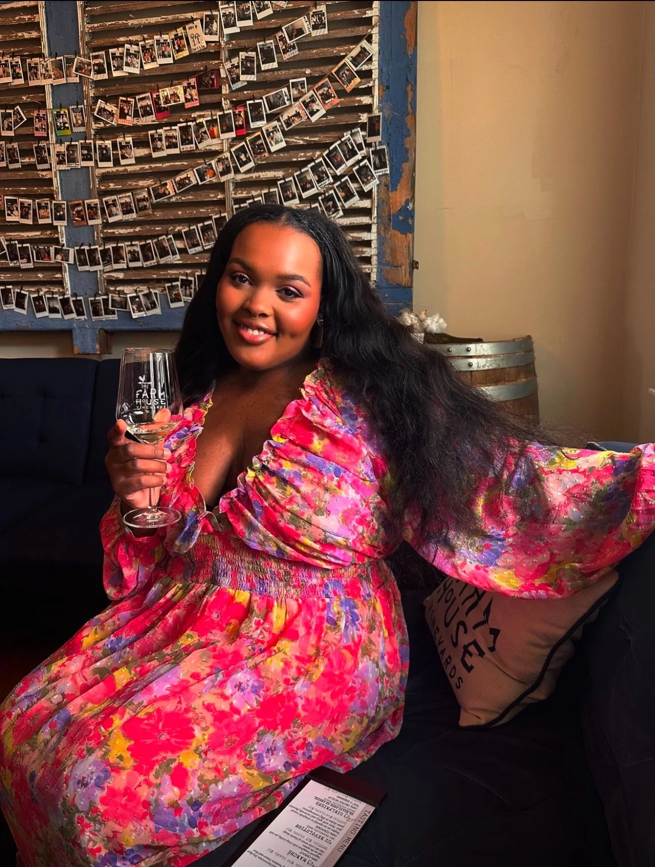 A woman with long dark hair, wearing a colorful pink floral dress, sitting on a dark sofa, holding a wine glass, smiling at the camera, in a cozy setting decorated with a wall of Polaroid photos and a wooden barrel.