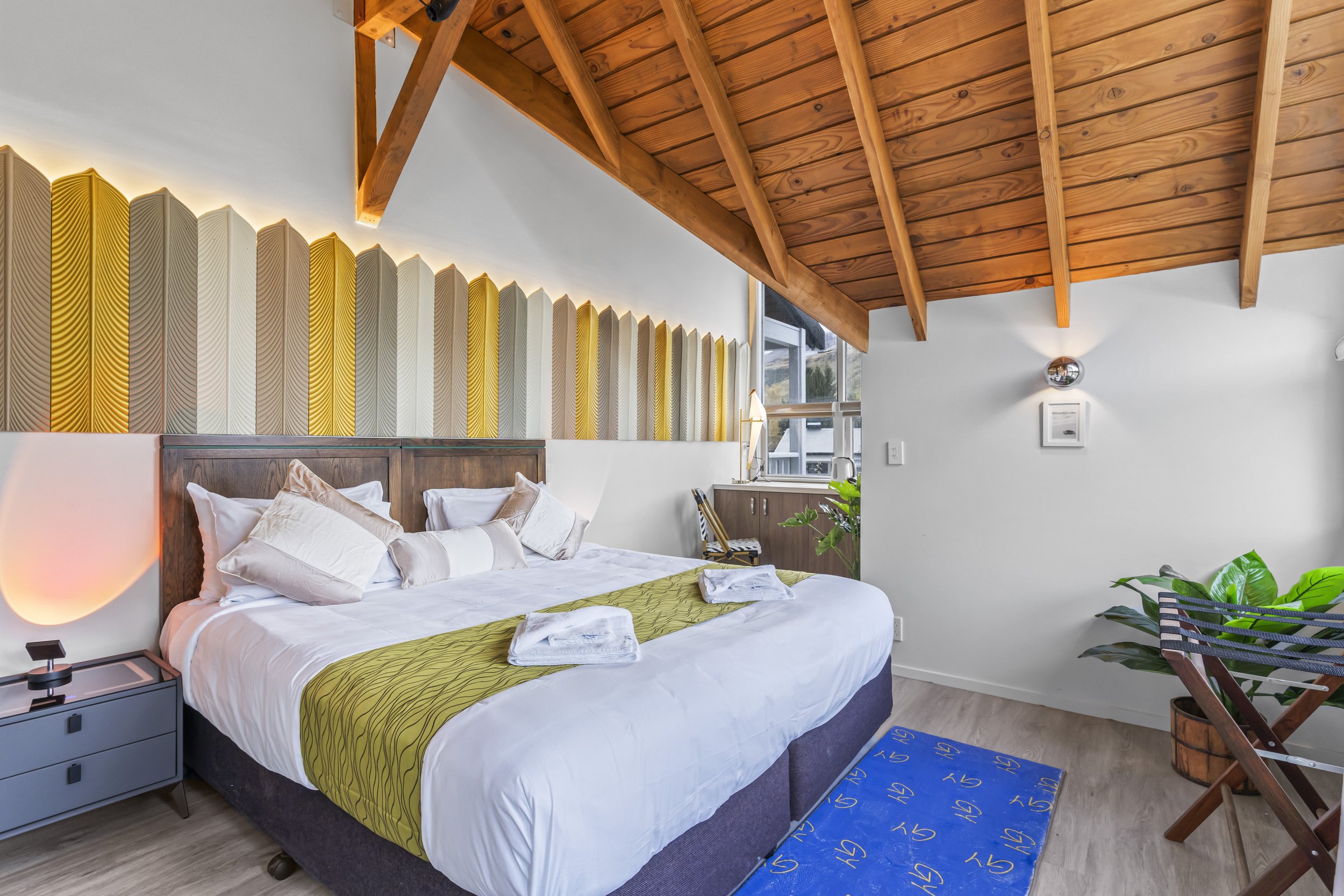 Hotel room with bed, decorative wall art, wooden ceiling, potted plant, and furniture.
