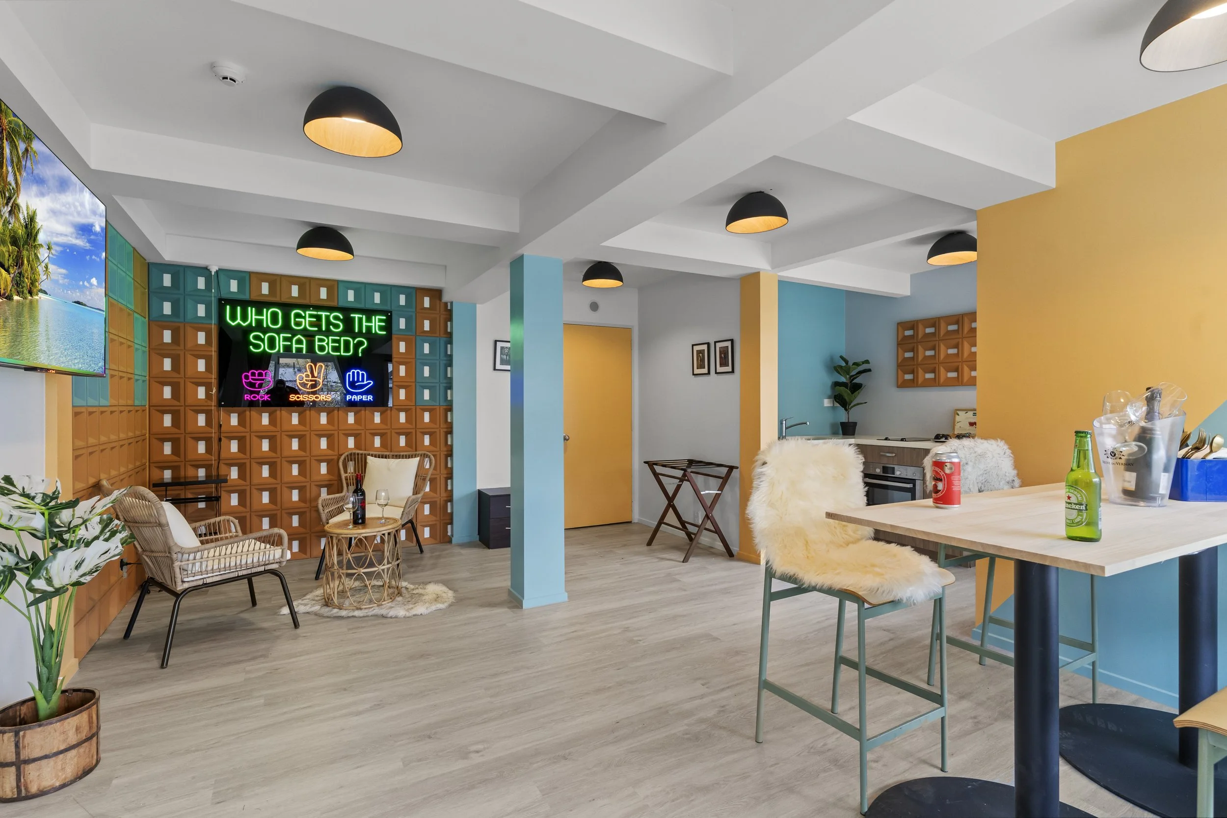 Colorful modern indoor space with yellow and blue walls, a camp stove setup, a wooden bar table with drinks, a TV on the wall, and a neon sign asking about who gets the sofa bed, featuring game prompts for rock, scissors, paper.