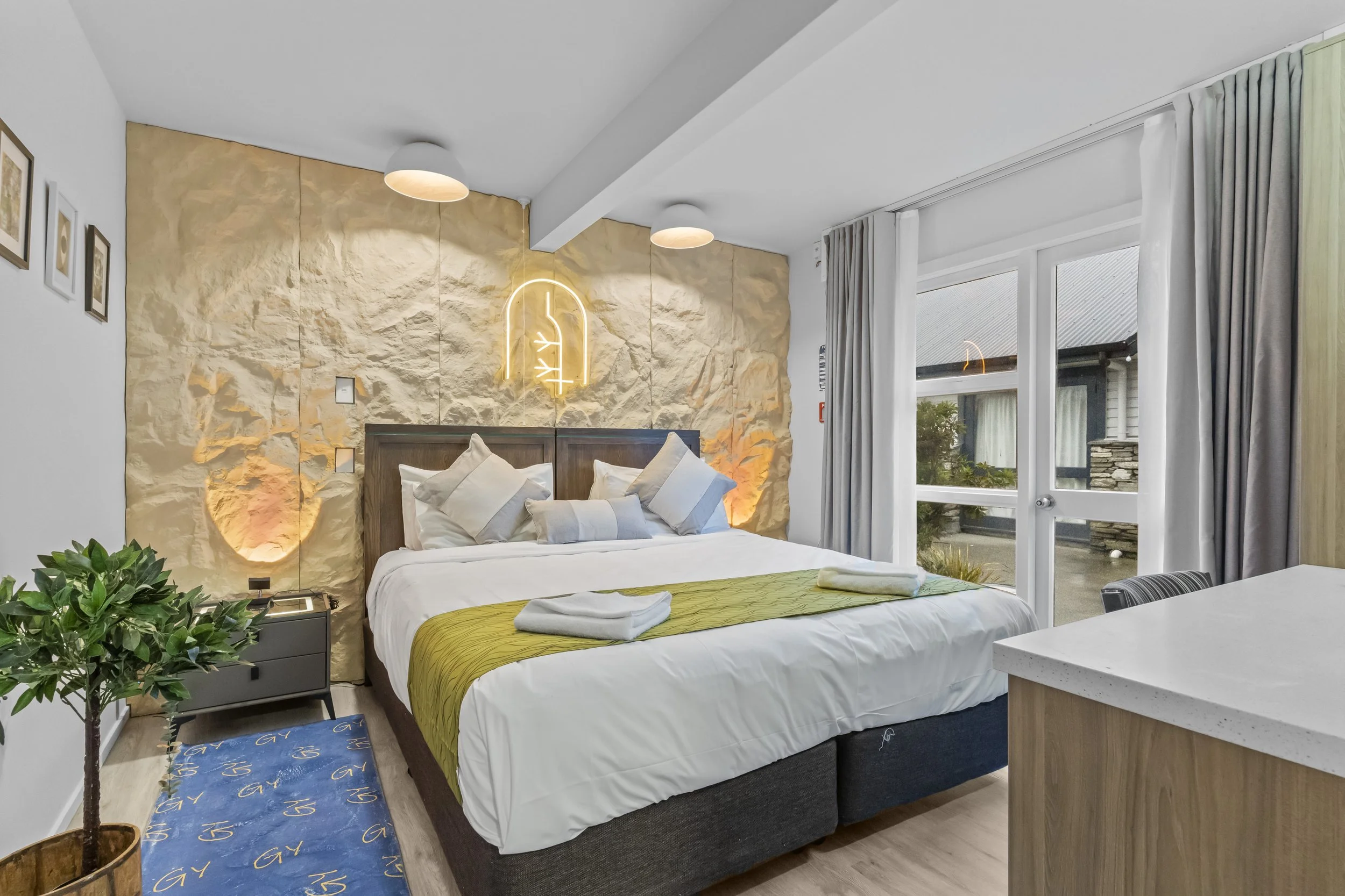 Modern hotel room with a large bed, decorated with white and beige pillows, a yellow throw, and towels. The room features a textured stone accent wall with a neon light art piece, a nightstand with a plant, and large windows with curtains showing an 