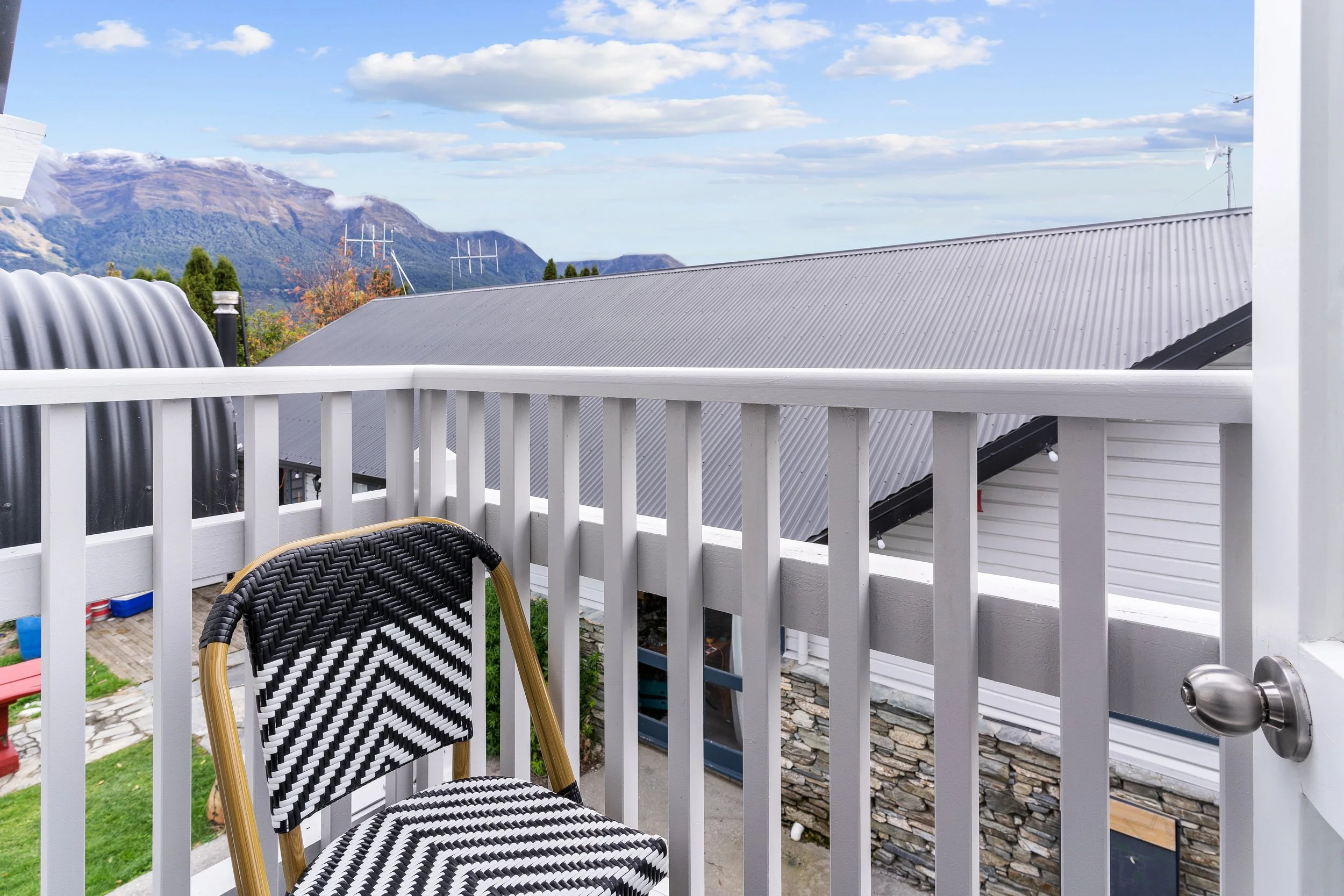 View from a balcony with a black and white woven chair, white railing, and neighboring rooftop with mountains and a partly cloudy sky in the background.