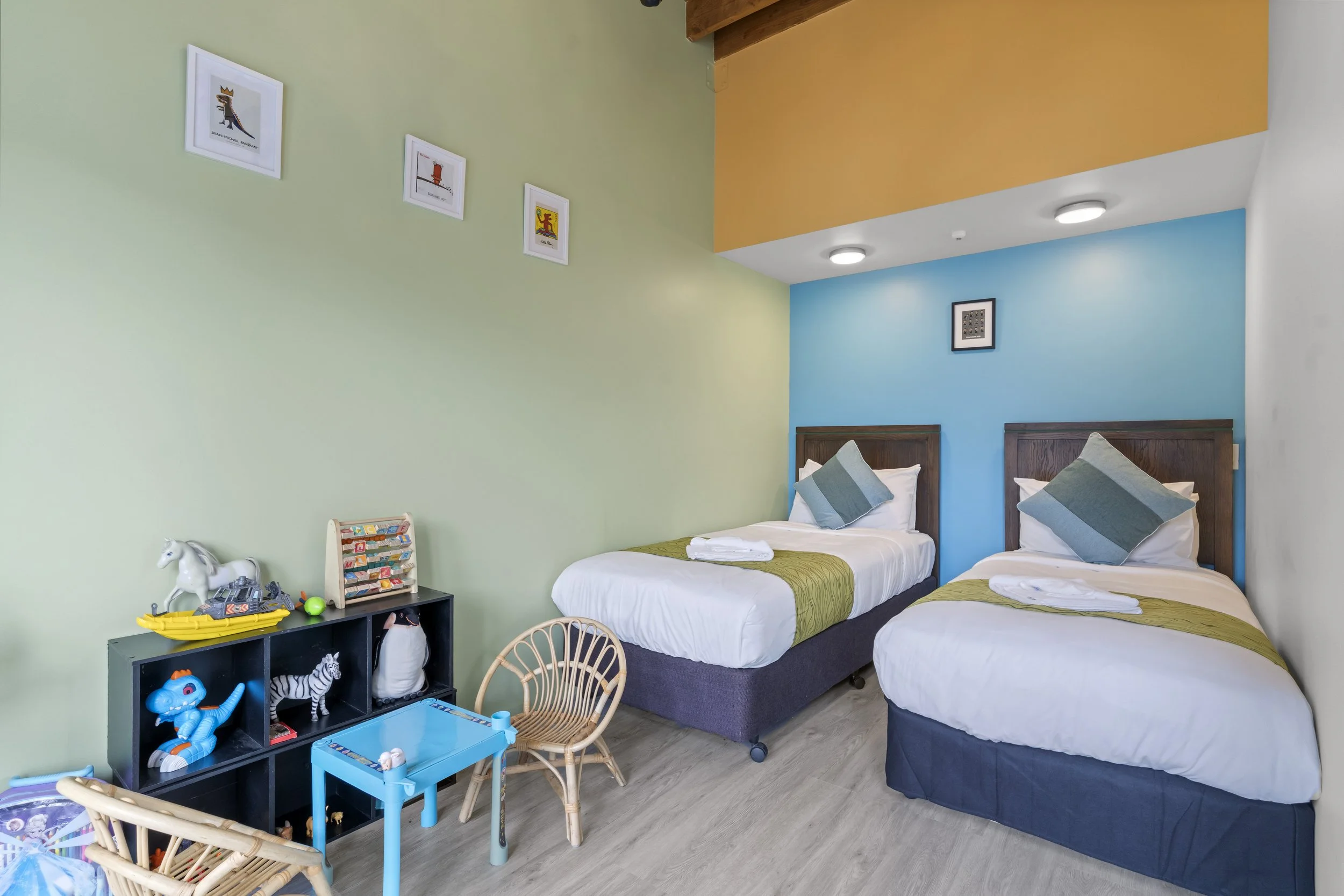 Two single beds with wooden headboards, each with white bedding, grey and green pillows, and folded towels. A blue wall behind the beds and a light green wall to the side with framed artwork. A children's toy shelf with various toys, a small blue tab