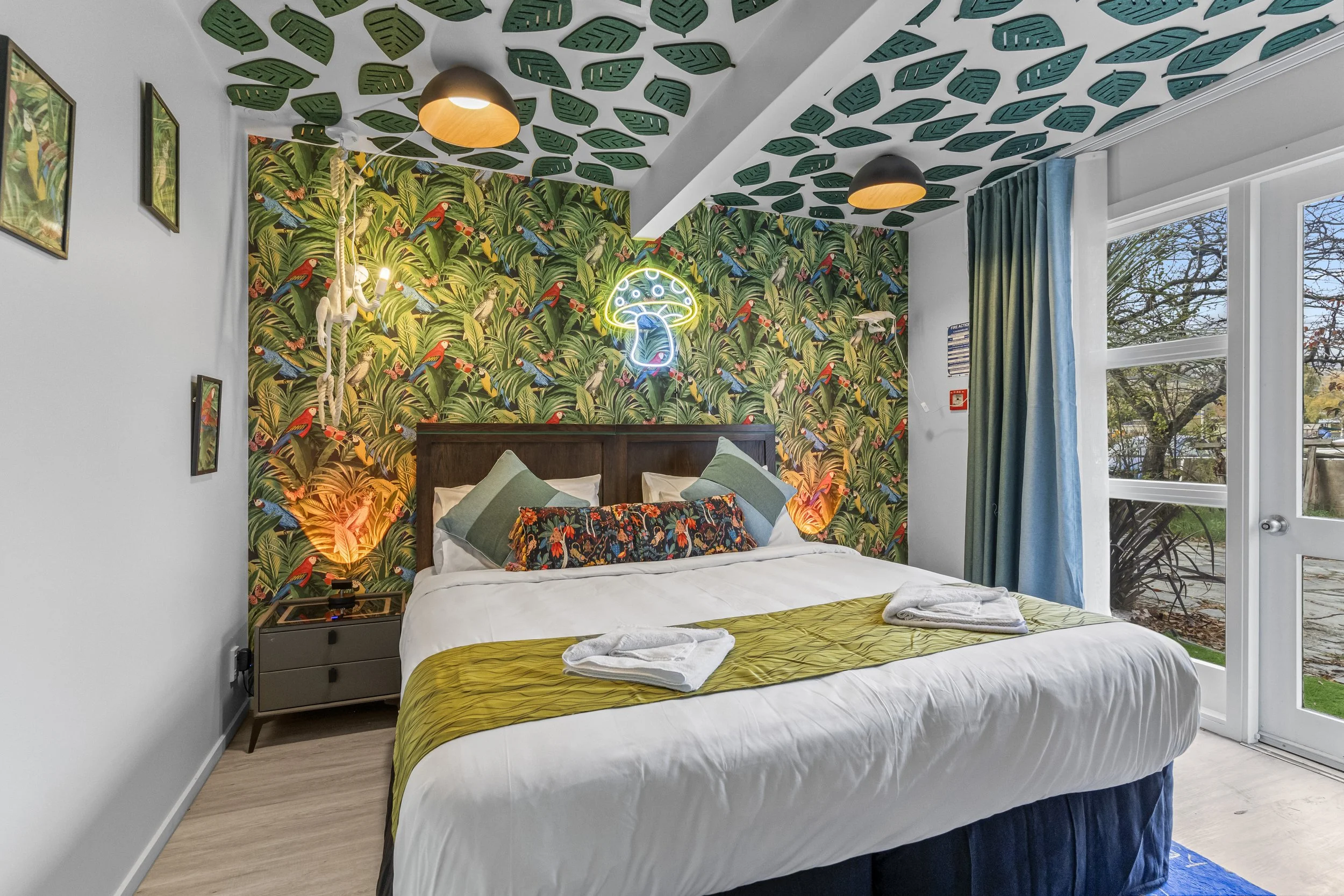 A bedroom featuring a tropical-themed wallpaper with lush greenery and colorful birds behind the bed. The bed has white bedding, green and floral pillows, and a yellow-green throw. Neon sign of a mushroom is on the wall. The ceiling has leaf-patterne