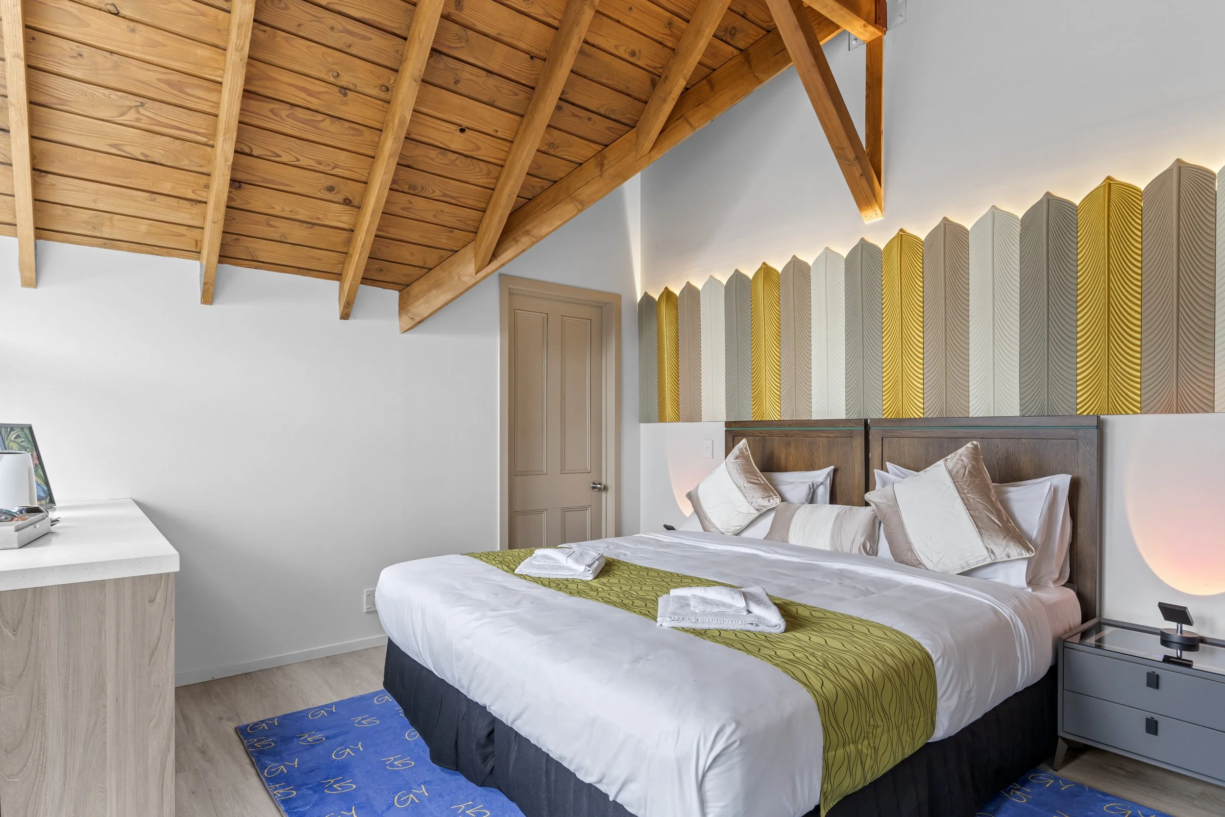 A neatly made hotel bed with white and beige pillows, a yellow-green runner, and two towel swans on top. There is a wooden bed frame and a decorative wall with yellow, white, and gray vertical panels behind the headboard. A bedside table with a lamp 