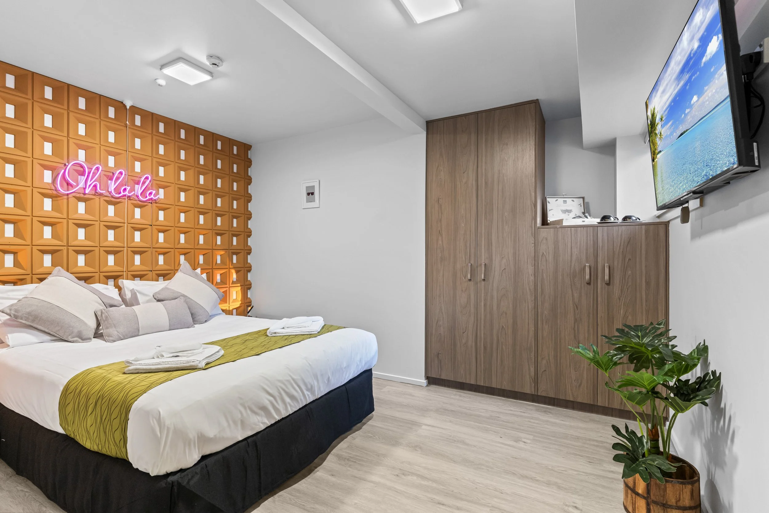 Modern hotel room with a bed, decorative orange headboard with a pink neon 'Oh La La' sign, a flat-screen TV, wooden wardrobe, potted plant, and white walls.