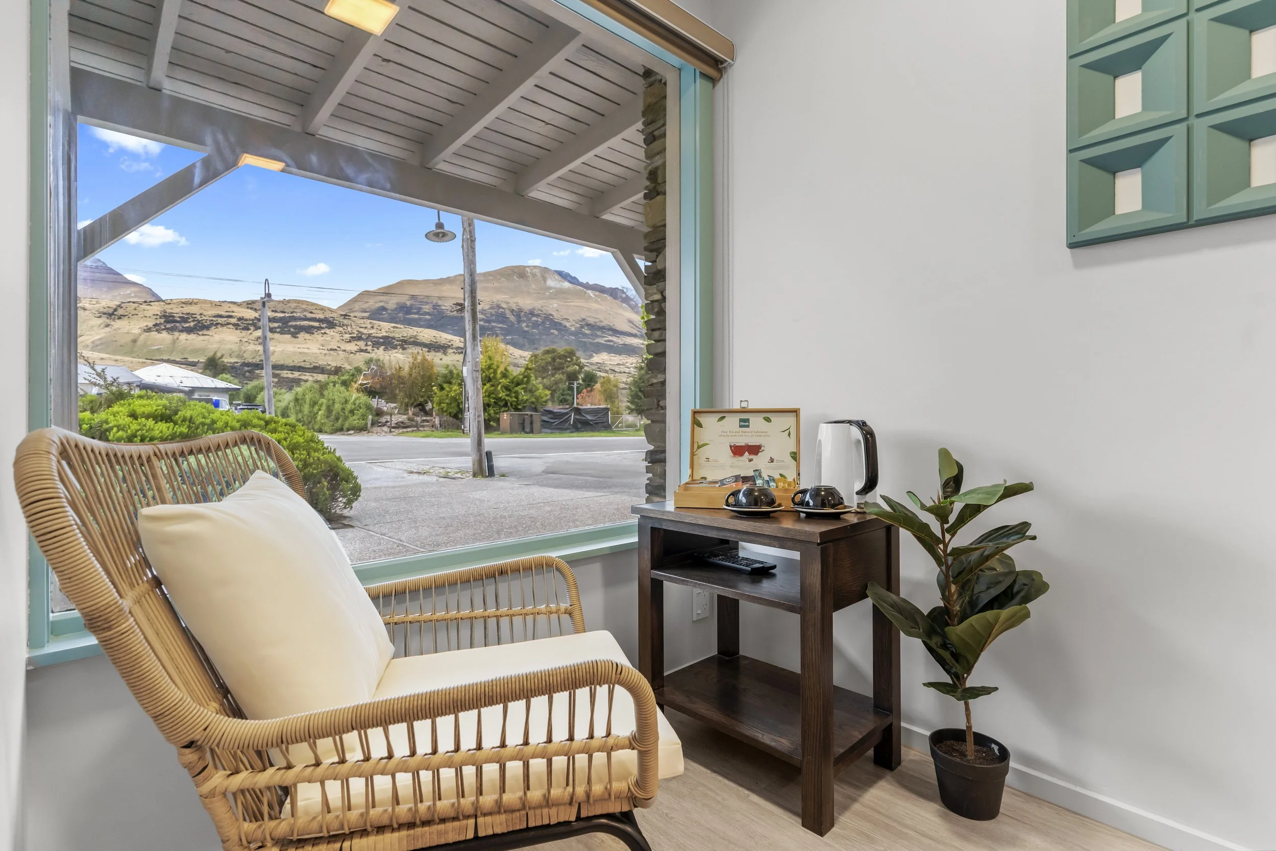 Interior of a cozy room with a rattan armchair, a small wooden table with a tea set and kettle, a potted plant, and large window showing a view of hills and a street outside.