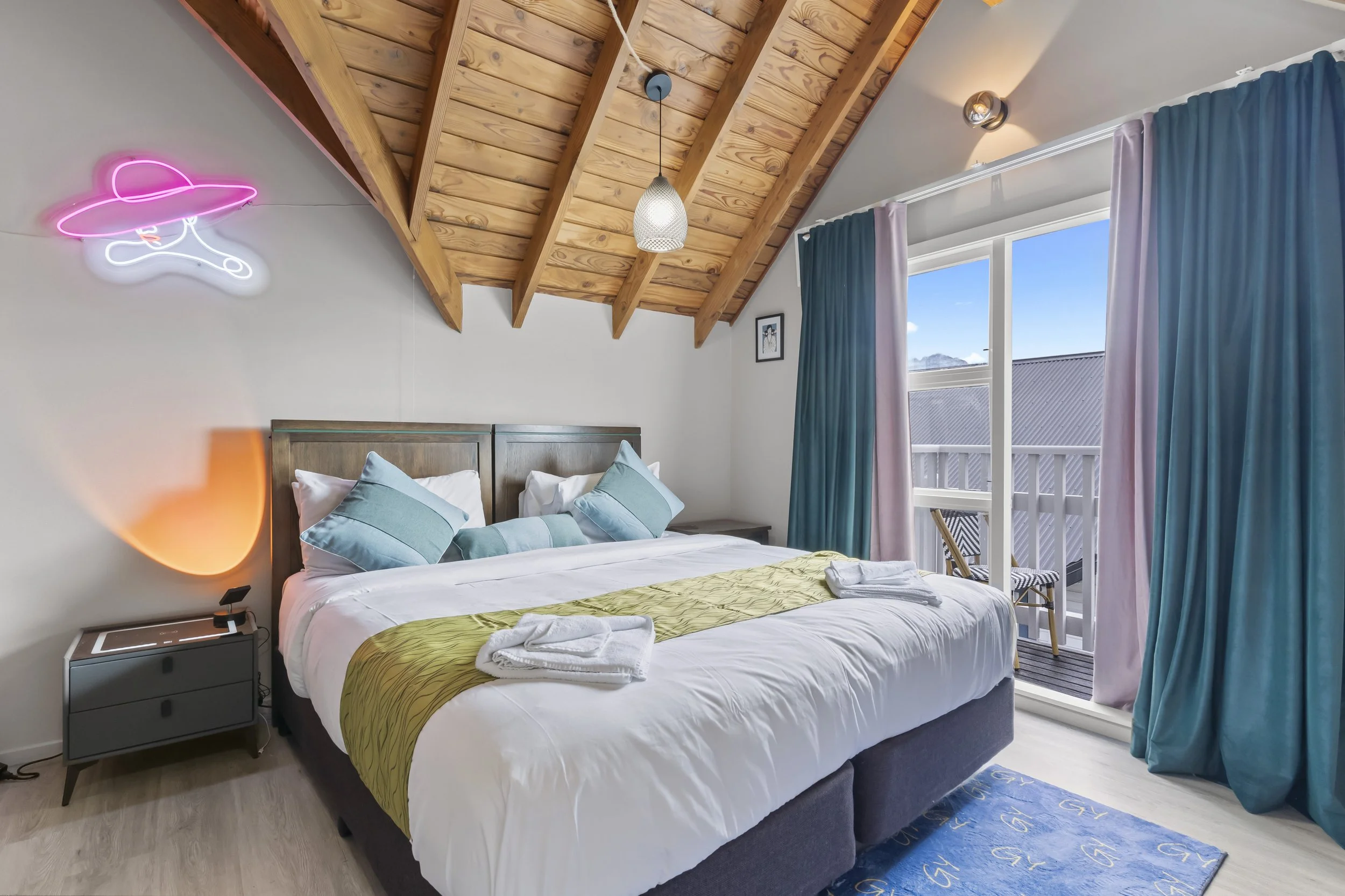 A cozy bedroom with a large bed, blue pillows, a yellow bed runner, and white bedding, next to a small nightstand with a phone, overlooking a balcony with blue curtains and a view of the sky.