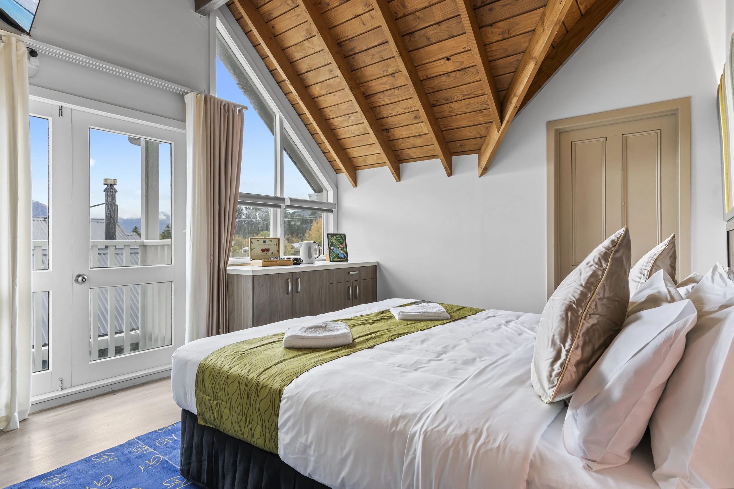 A cozy bedroom with a slanted wooden ceiling, a large bed with white sheets and pillows, a green bed runner, and a window letting in natural light. There are towels on the bed and a cabinet with decorative items near the window.