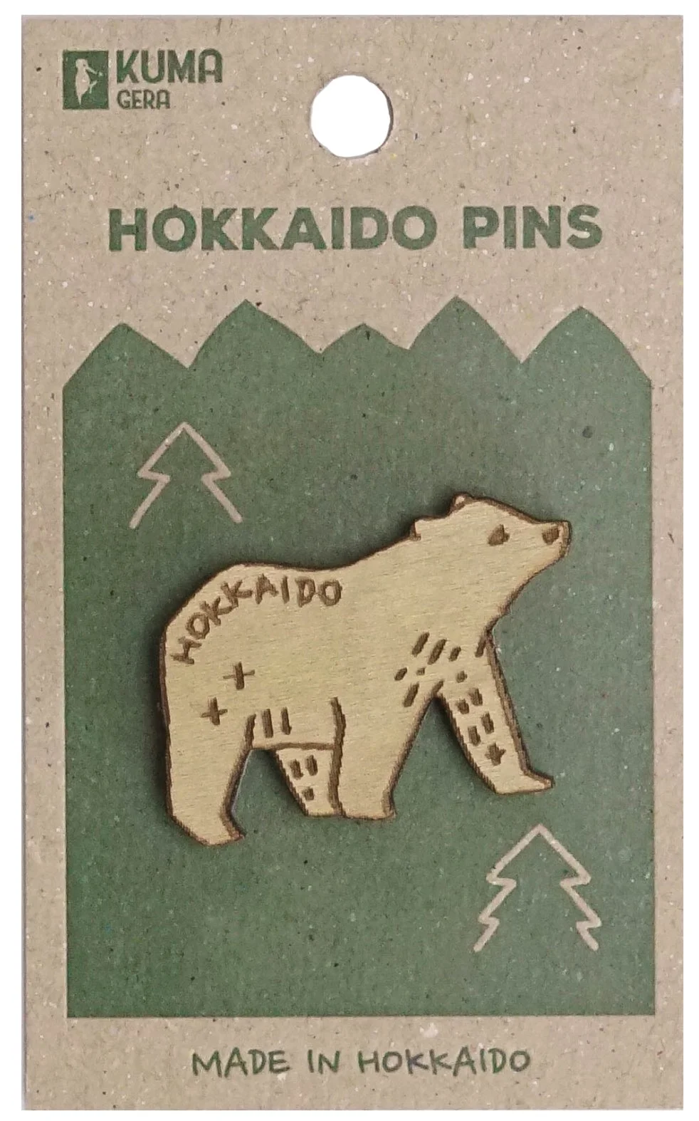 Wooden pin in the shape of a bear with the word 'Hokkaido' engraved on it, mounted on green packaging labeled 'Hokkaido Pins' and 'Made in Hokkaido.' The packaging also features mountain peaks and lightning bolt symbols and a small logo of a bear in the top left corner.