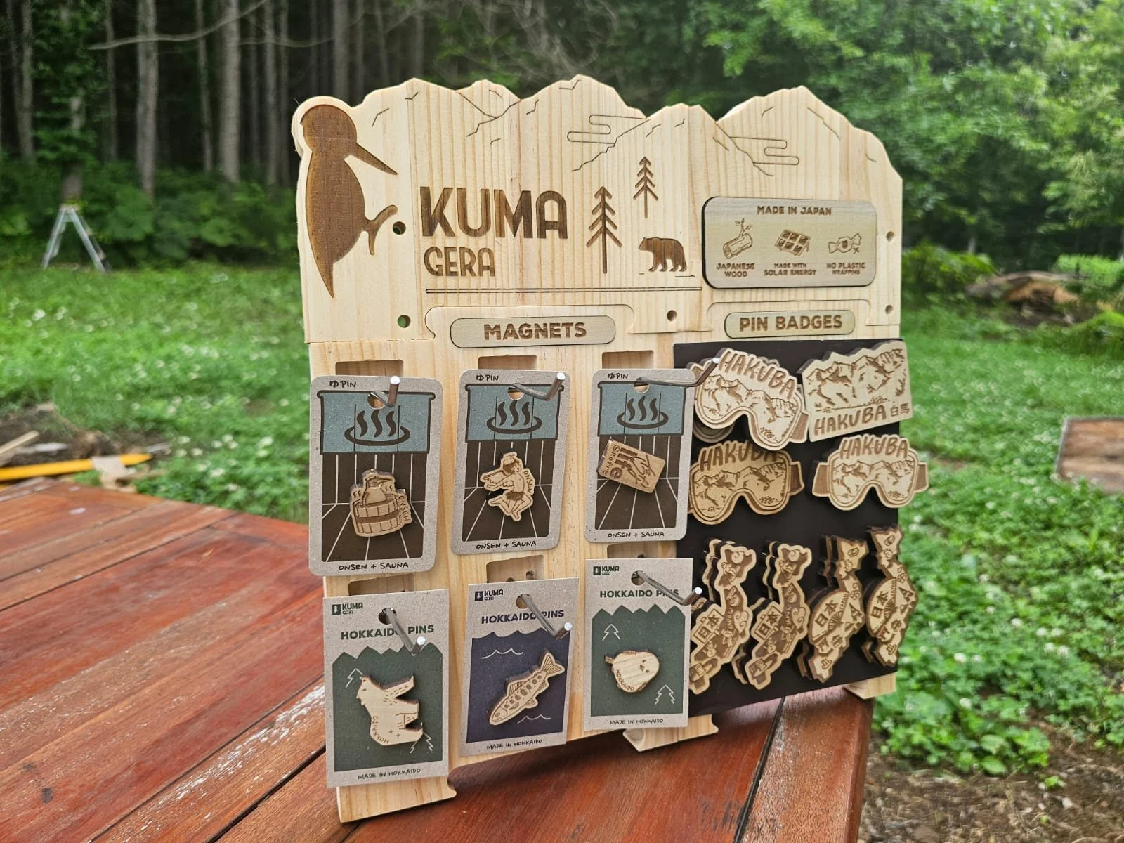 Wooden display board featuring magnets and pin badges related to Hokkaido, Japan, with images of mountains, bears, and local icons, set outdoors in a grassy area.