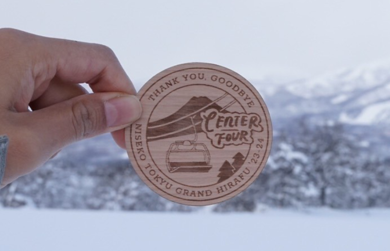 Hand holding a wooden token with text, mountains, and cable car illustration in background, snow-covered landscape.