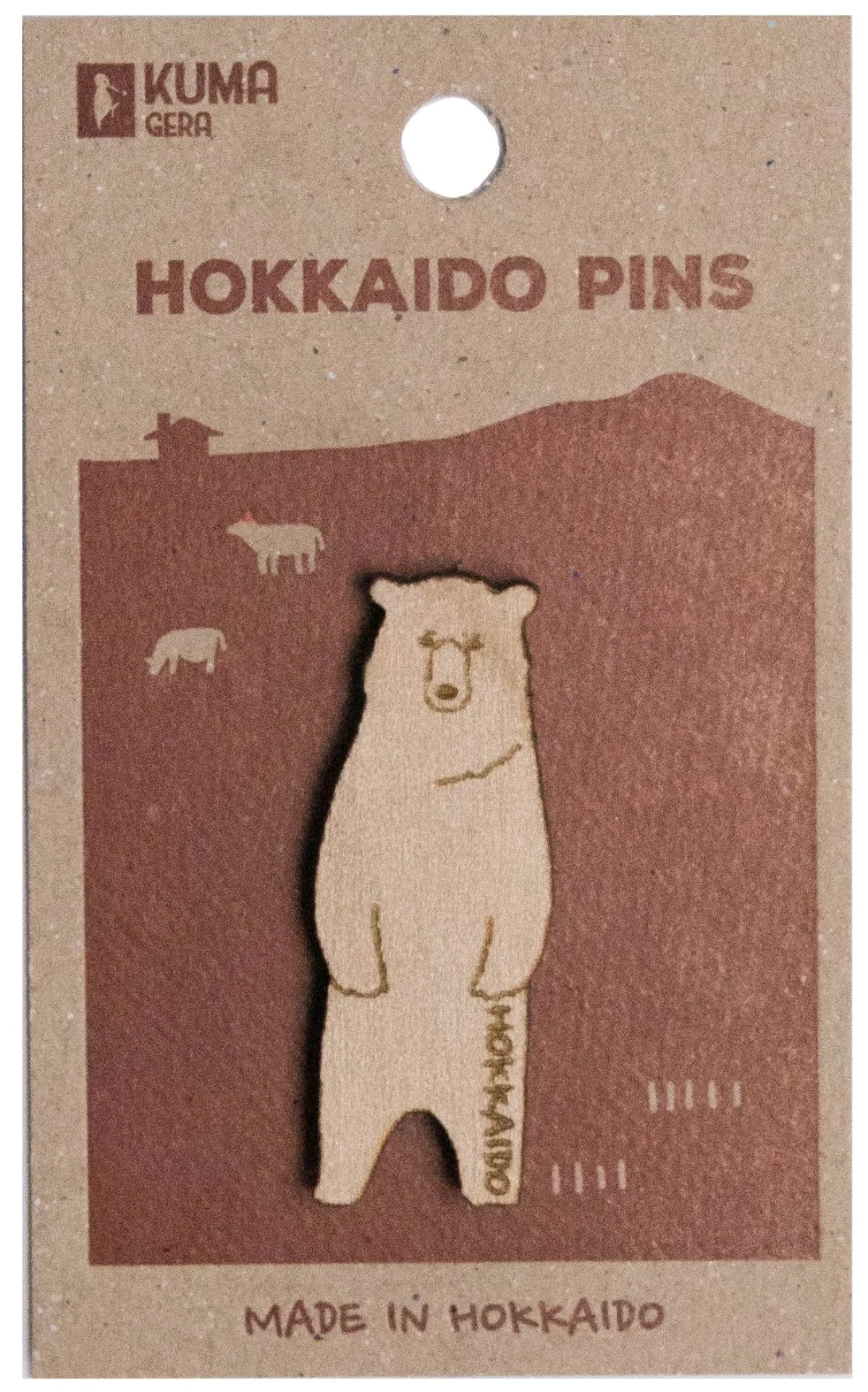 A Hokkaido-themed pin featuring a bear design on brown cardboard packaging with text indicating it is made in Hokkaido. The pin itself resembles a bear and includes small illustrations of animals and scenery typical of Hokkaido.