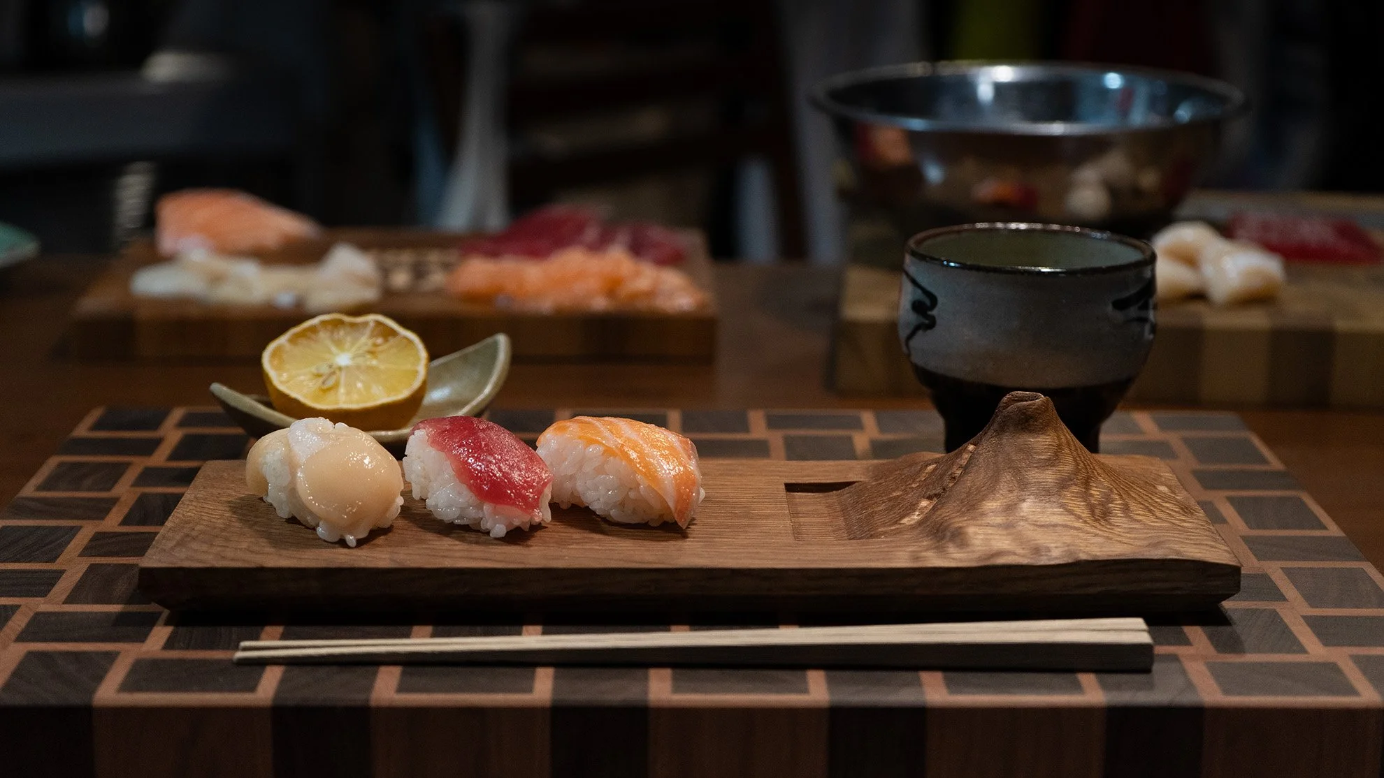 Assorted sushi pieces on a custom-made wooden platter with lemon slices, chopsticks, and a black tea cup on a patterned table.