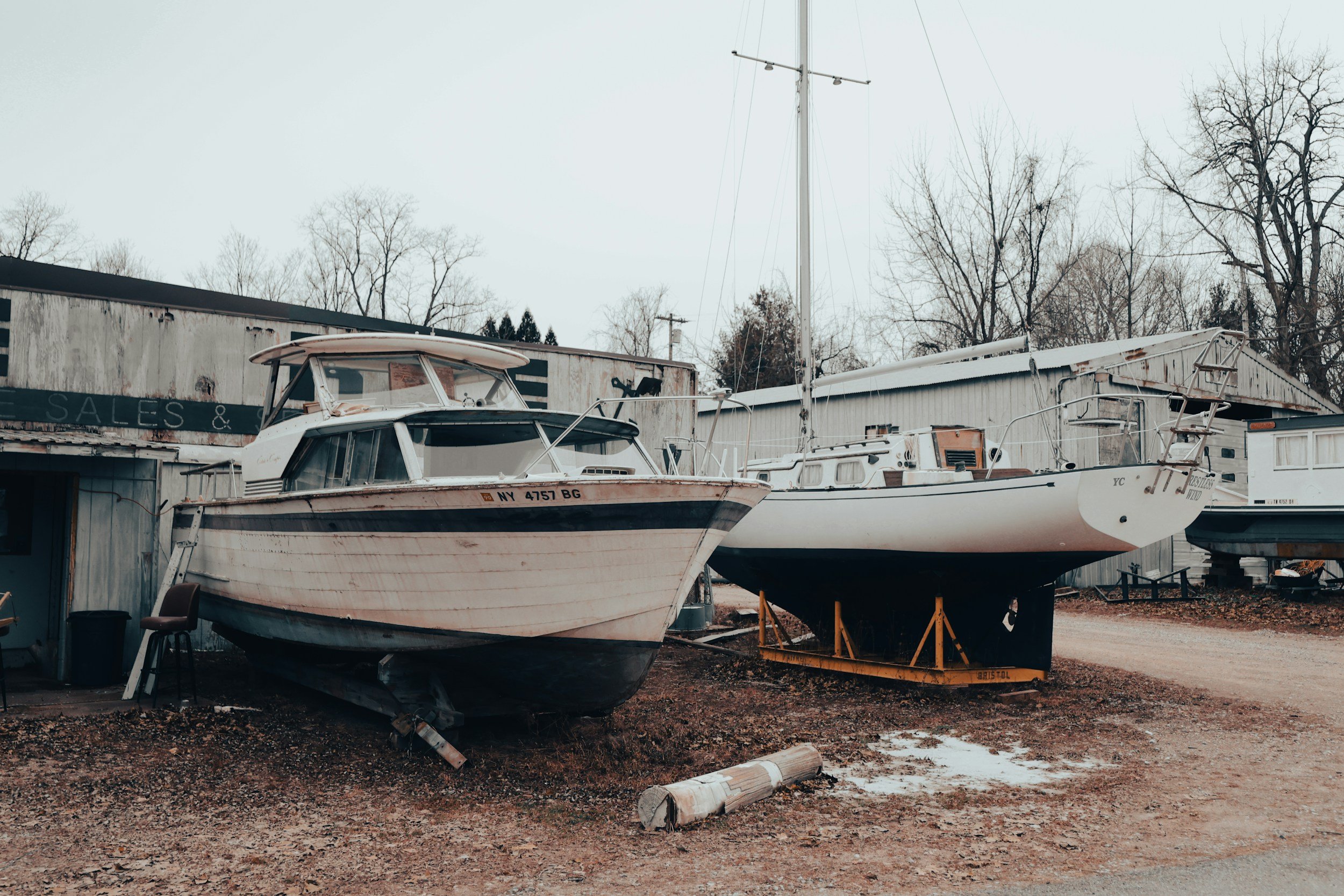 Two boats on land in a boatyard, one with a cabin and registration number NY 4757 BG, and the other is a sailboat. There are trees in the background and a weathered building.