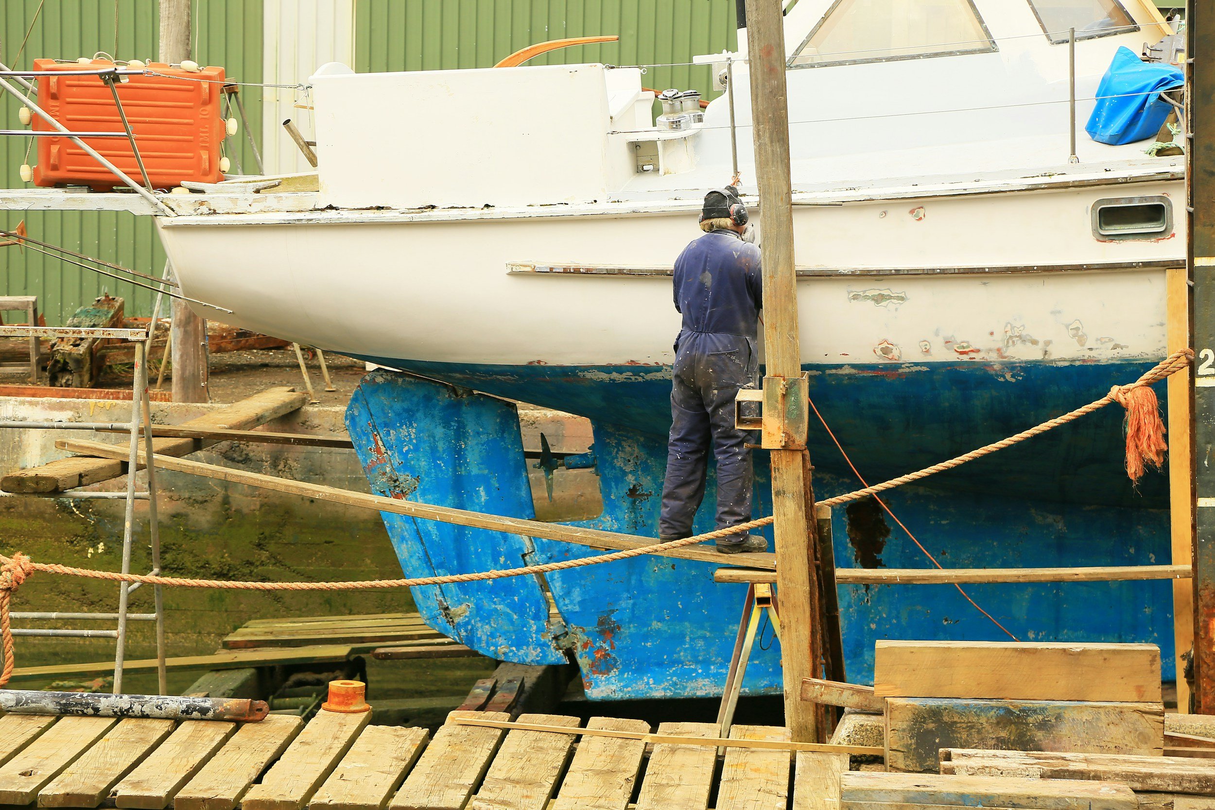 A person working on a boat on land, surrounded by wooden planks and scaffolding, with a green corrugated metal building in the background.
