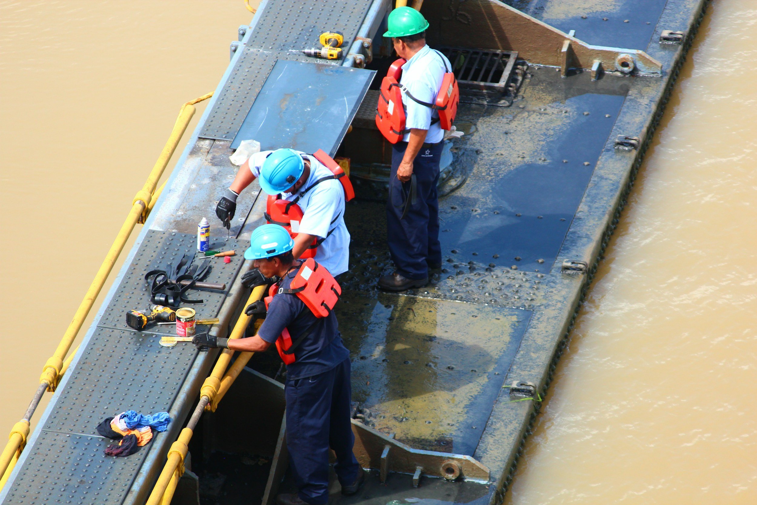 Three workers wearing protective helmets and life vests on a floating platform working on equipment near a body of muddy water.