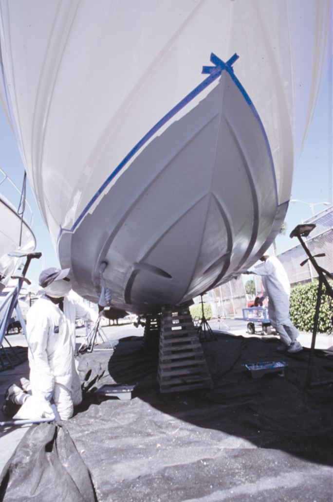 Two workers in protective clothing and gloves inspecting the hull of a large boat elevated on wooden supports at a boatyard, with tools and equipment nearby.