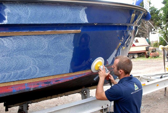 Man polishing the hull of a blue boat on a trailer at a marina or boatyard.