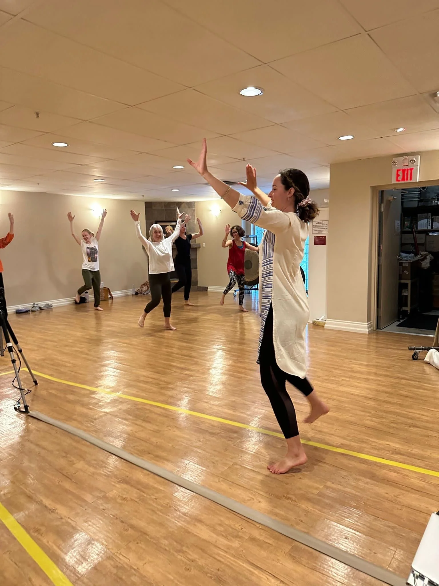A group of women practicing dancing or yoga in a studio with a instructor leading, all with arms raised in a pose, standing on a wooden floor.