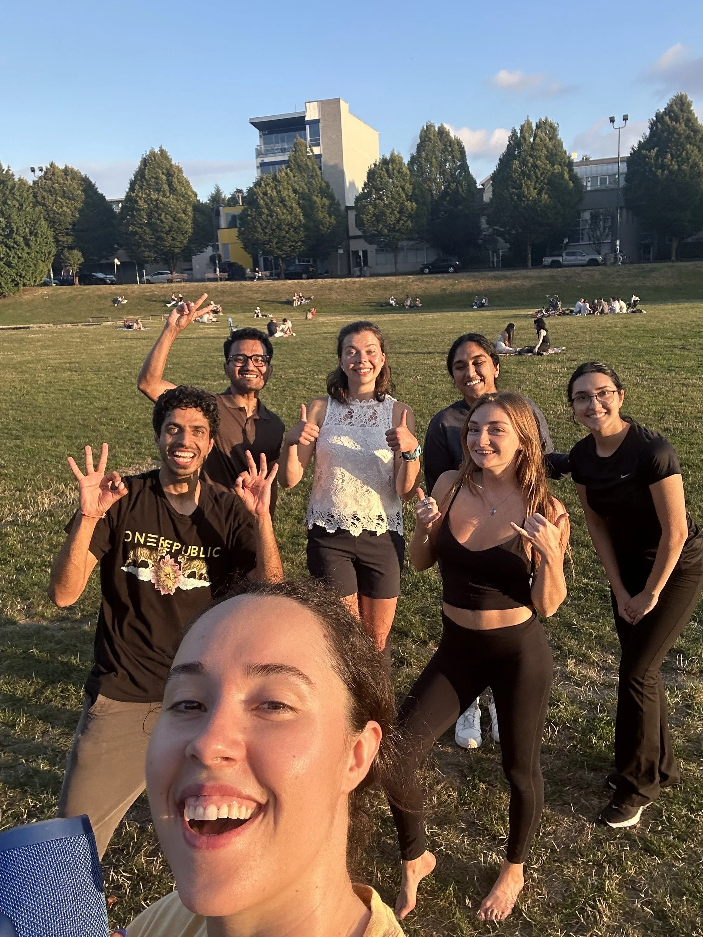 Group of six friends smiling and posing for a selfie in a grassy park during the daytime, with a background of trees and buildings.