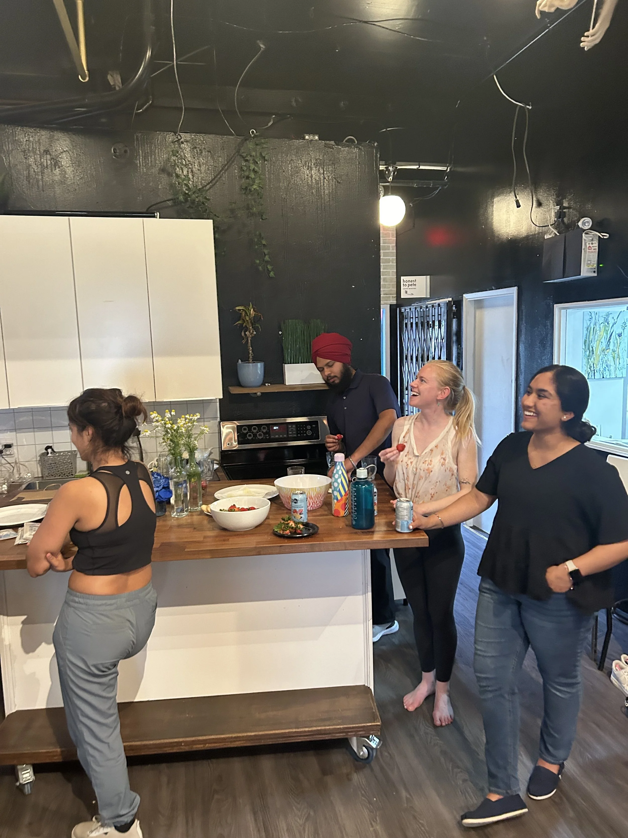 Group of friends enjoying a casual gathering in a modern kitchen, with some smiling and holding snacks, and one person looking at their phone.