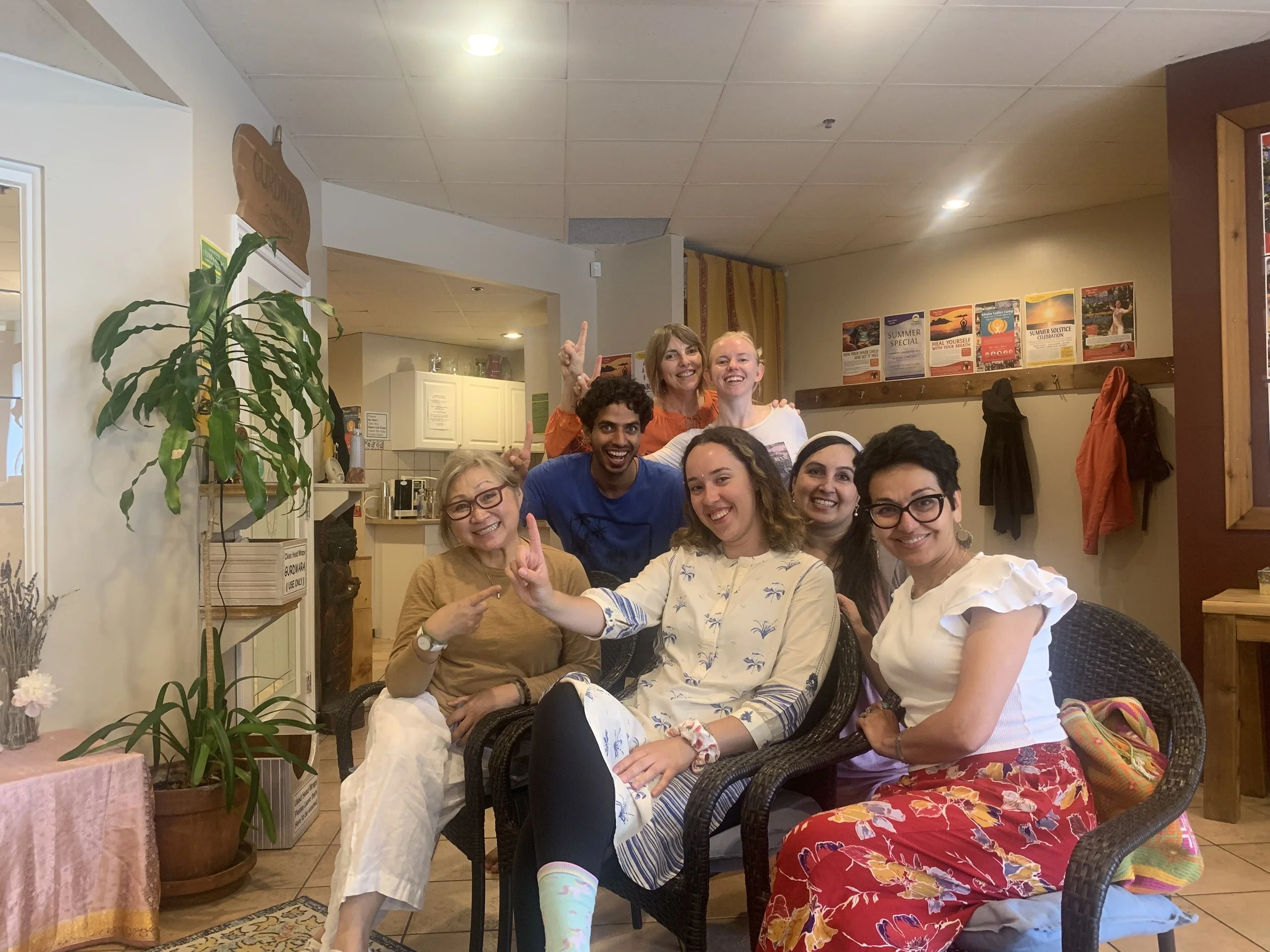 Group of eight diverse women and one man smiling and posing together in a cozy indoor setting, with some seated and others standing behind, all dressed in casual clothing.