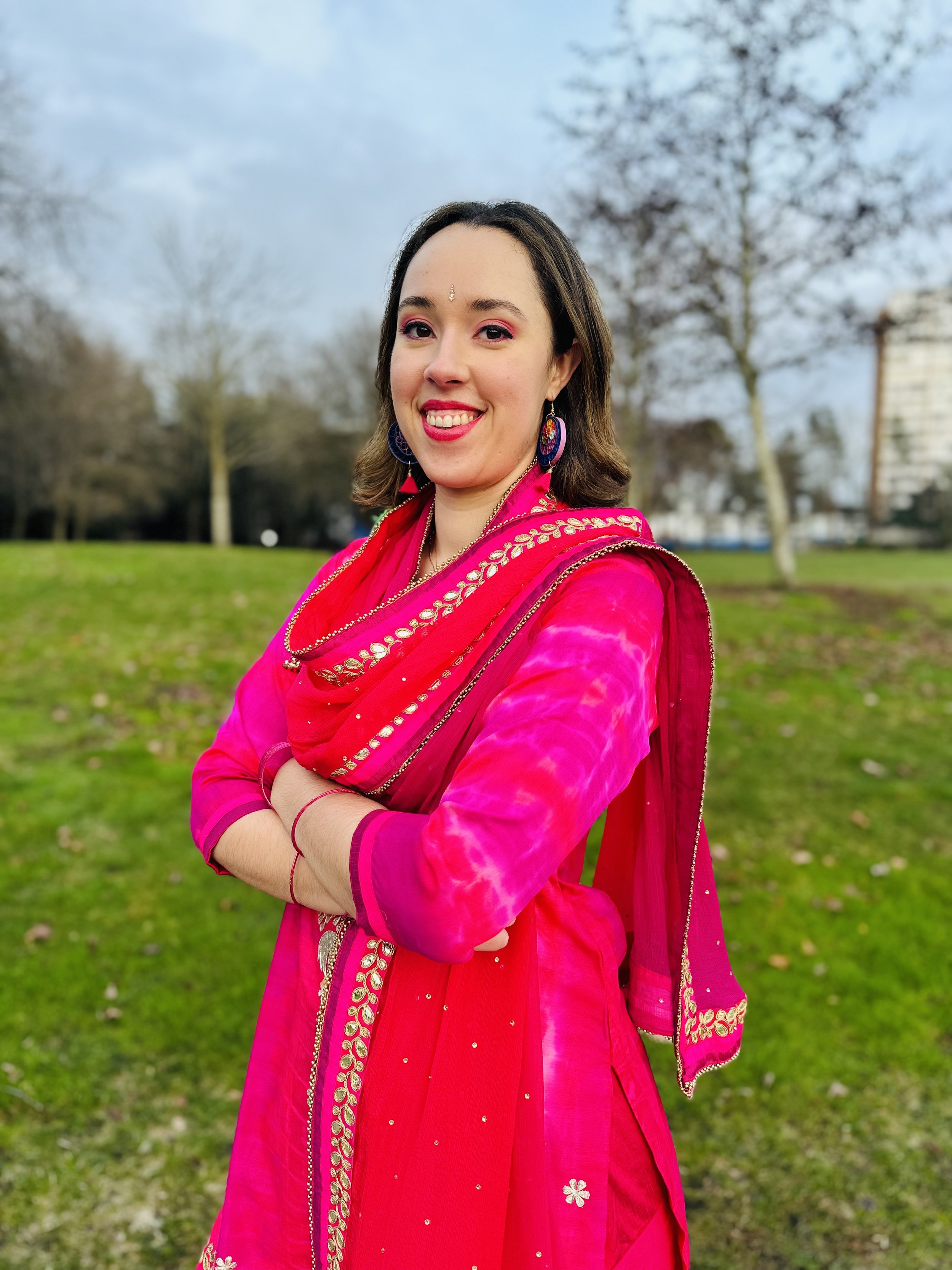 A woman in a bright pink traditional dress with gold accents, standing outdoors on a grassy park with trees and cloudy sky in the background.