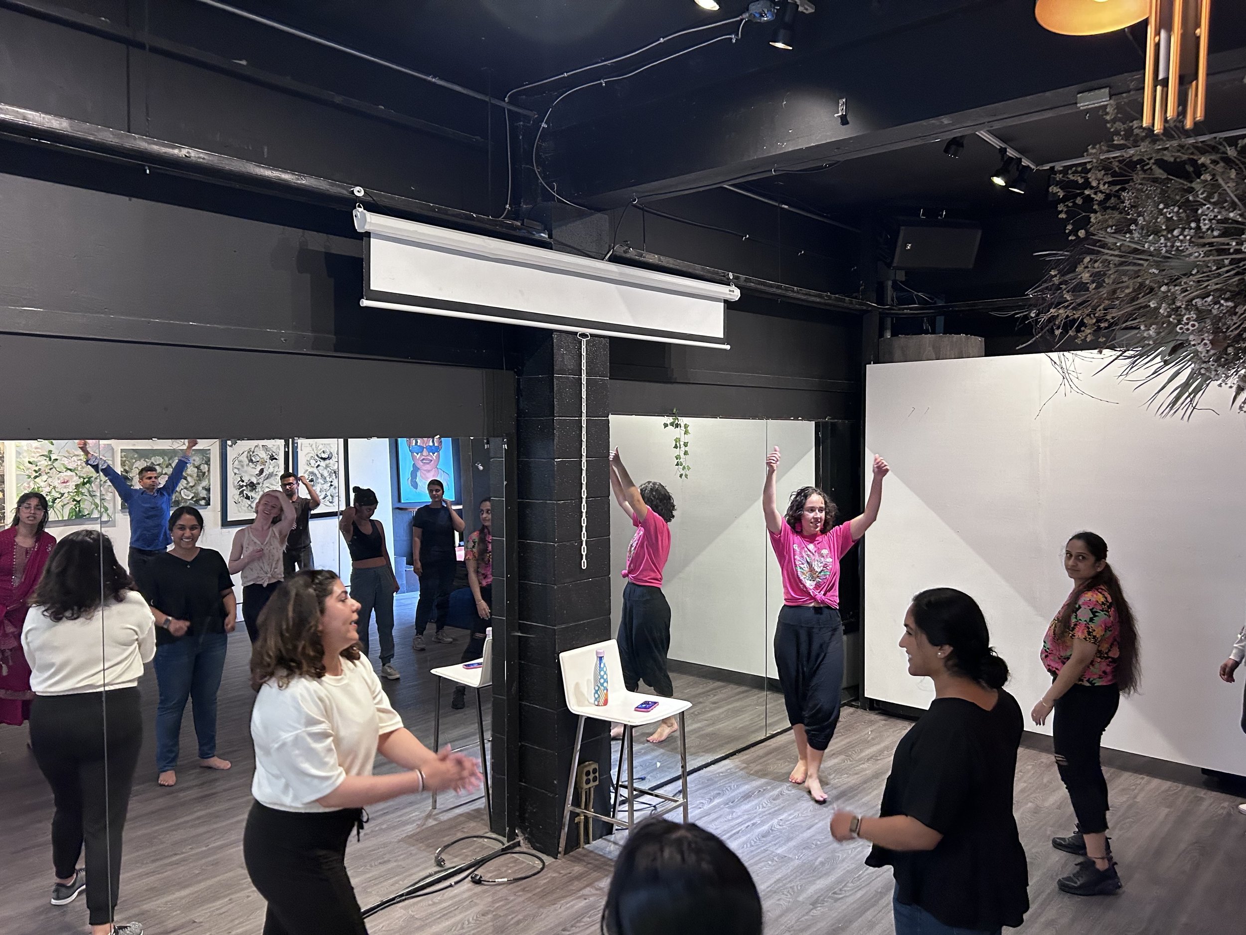 A group of people participating in a dance or movement class in a studio with reflective mirrors and artwork on the walls, some wearing pink shirts and others in different attire, appear to be enjoying the activity.