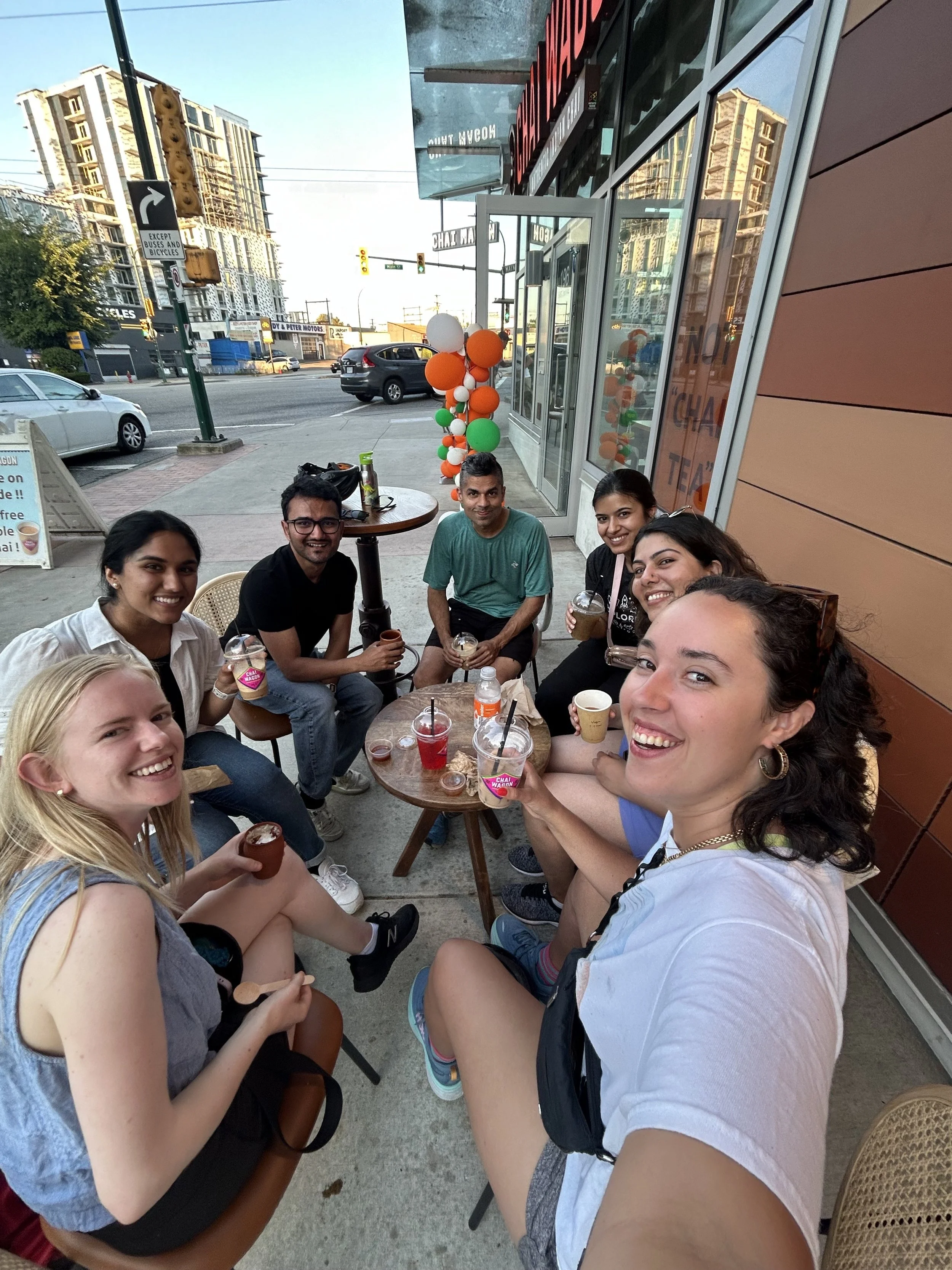 A group of seven friends sitting outside a cafe, smiling at the camera, with colorful balloons and drinks on the table, on a city street with buildings and cars in the background.