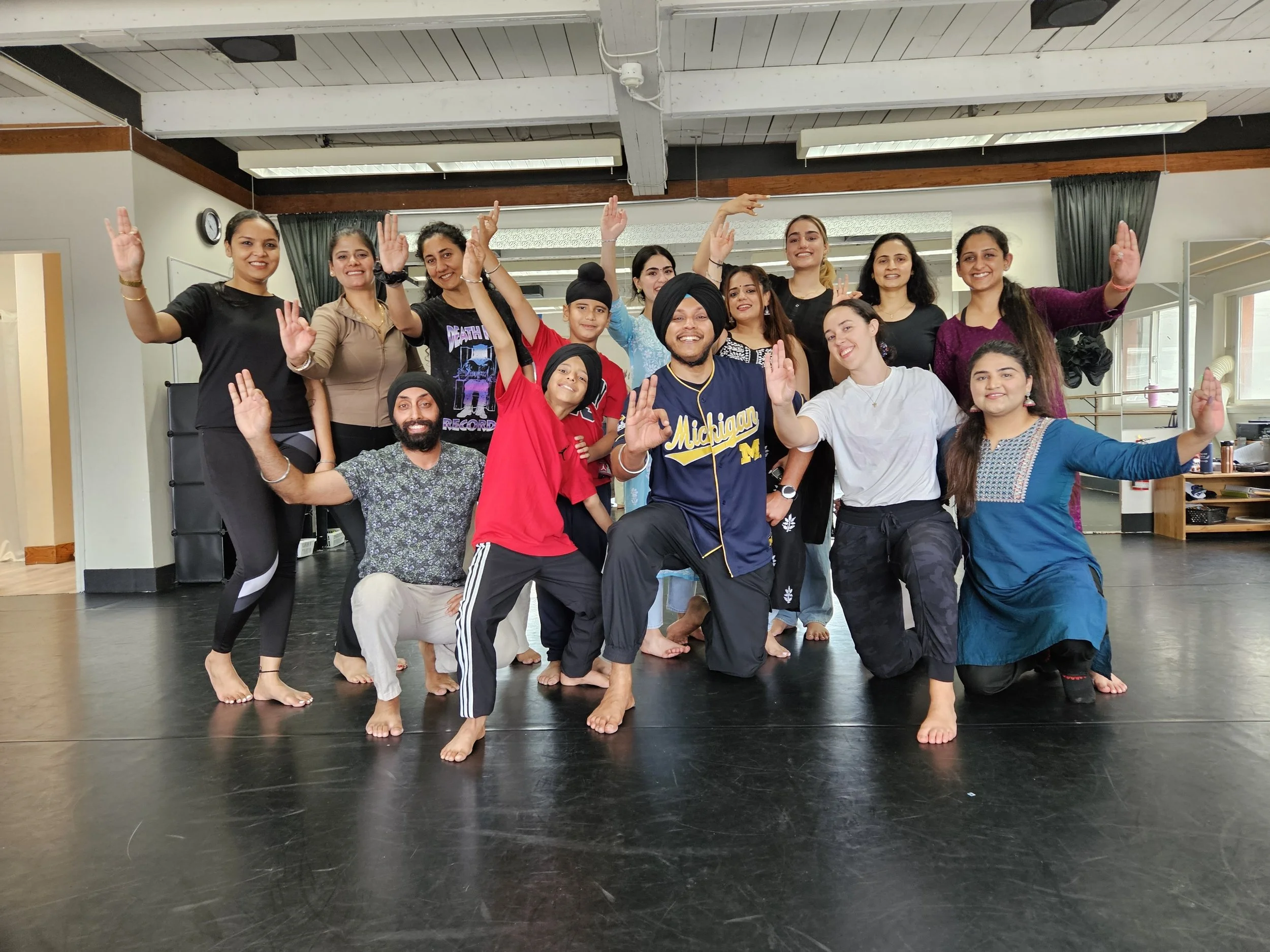 Group of people, mostly women and a few men, standing on a dance studio floor, smiling and posing with raised hands, some barefoot, in casual and traditional attire.