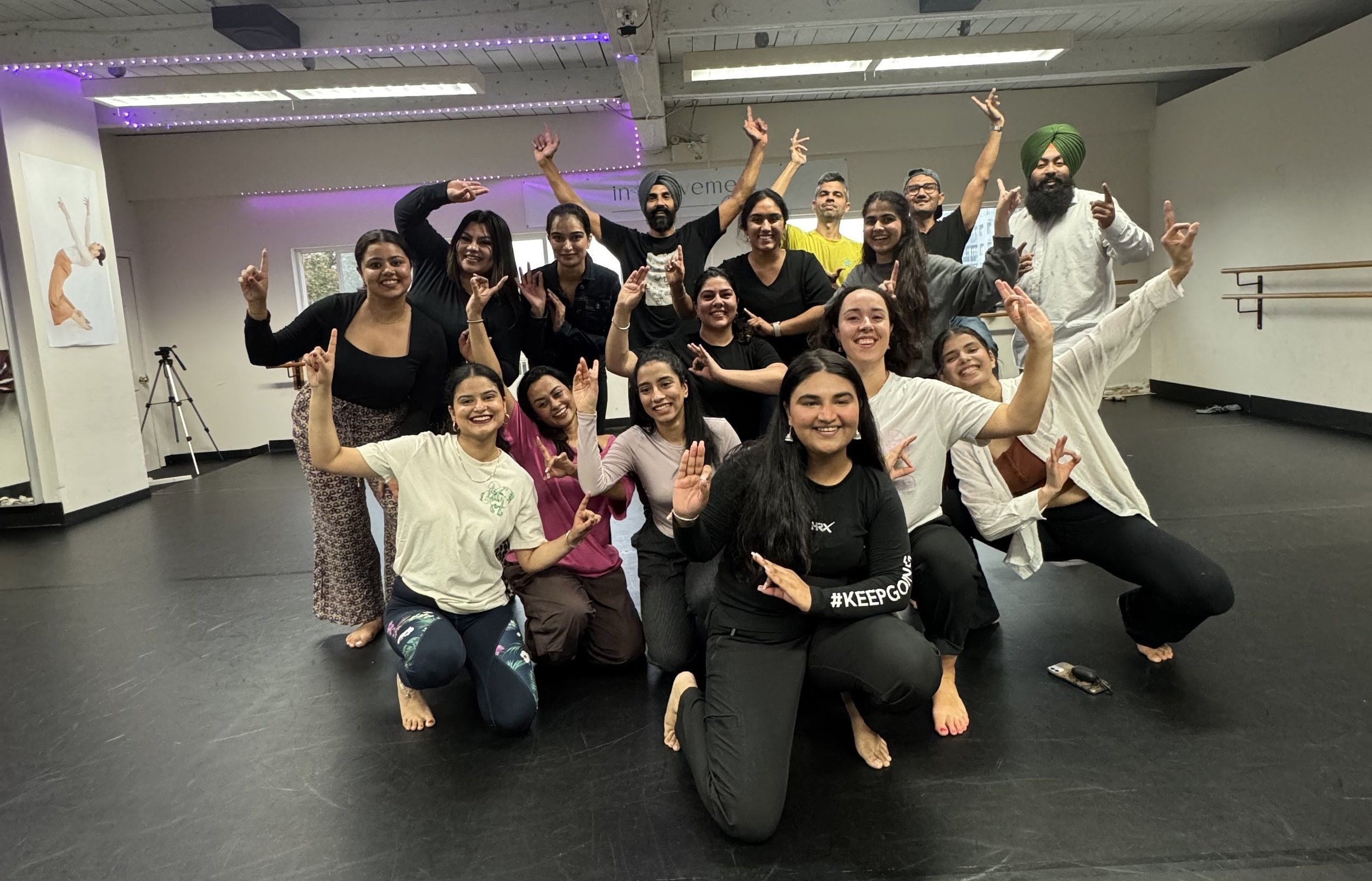 Group of women and men posing together in dance studio, smiling and making hand signs, some kneeling and some standing, with ballet barres and dance posters on the wall behind them.
